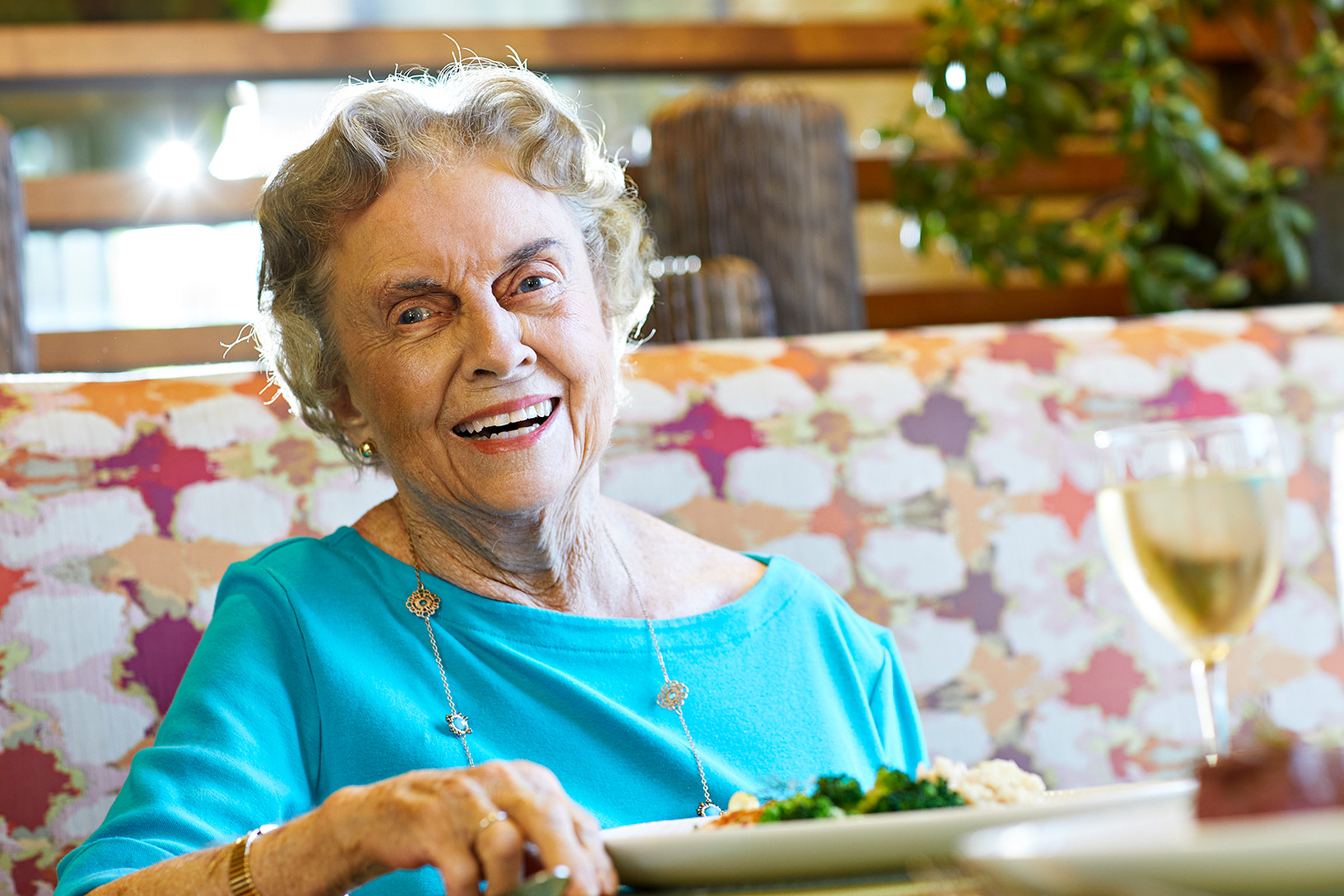 An elderly woman with short gray hair and a blue top smiles while sitting at a table with a plate of food and a glass of white wine, in a brightly lit restaurant.