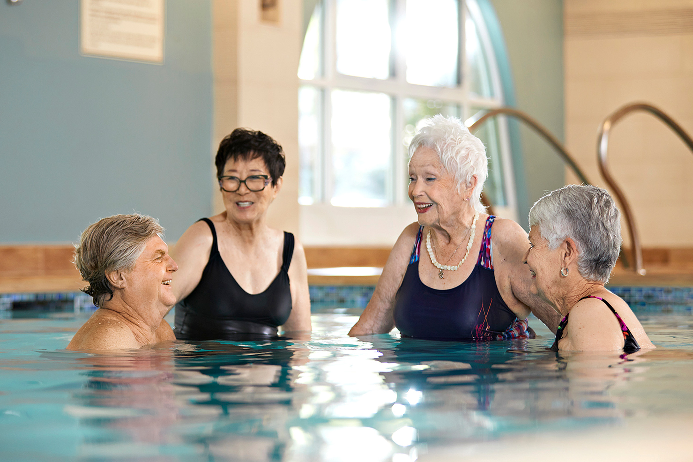 Four older women are standing together in a swimming pool, smiling and chatting. Sunlight comes through a large window in the background, creating a bright and cheerful atmosphere.