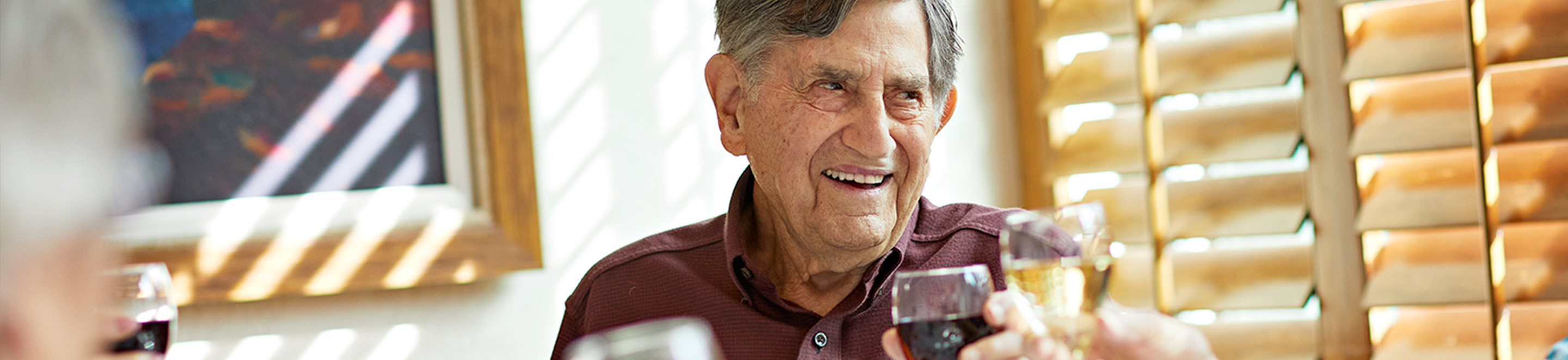 An older man smiles while holding a glass of wine, sitting at a table with others in a sunlit room with wooden blinds and a framed picture on the wall.