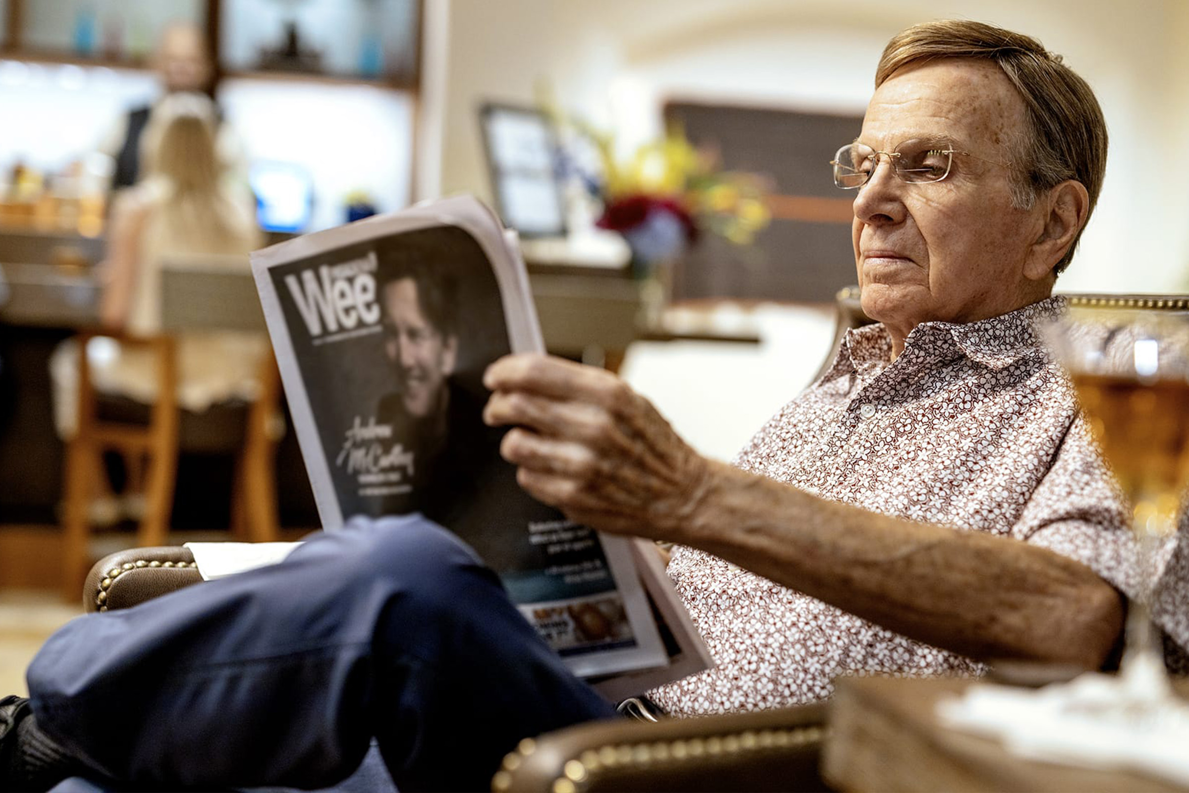 An elderly man wearing glasses and a patterned shirt sits comfortably in a chair, reading a magazine titled Wee in a well-lit room with blurred background figures and decor.