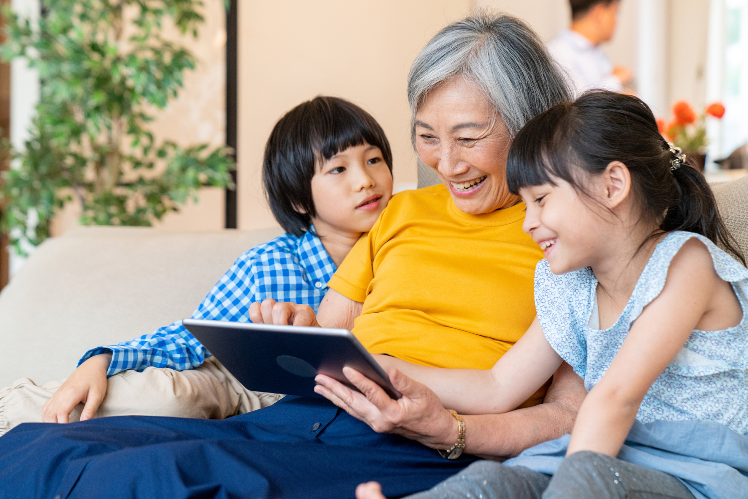 An older woman smiles while sitting on a couch with two children, a boy and a girl, as they look at a tablet together in a bright living room.