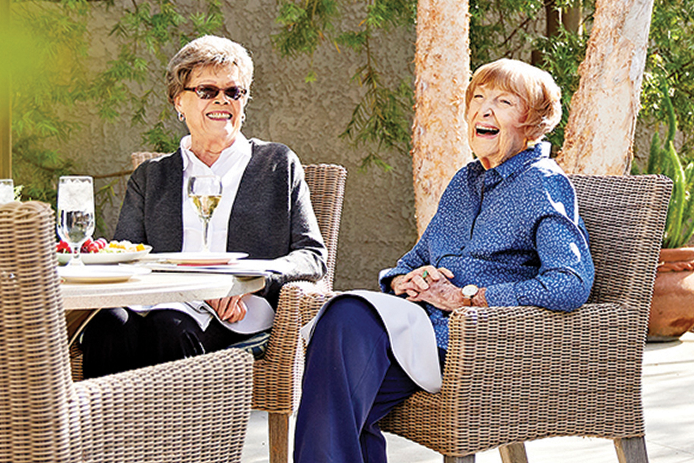 Two elderly women sit outside at a table, smiling and laughing together. They are enjoying drinks and food on a sunny day, surrounded by greenery.