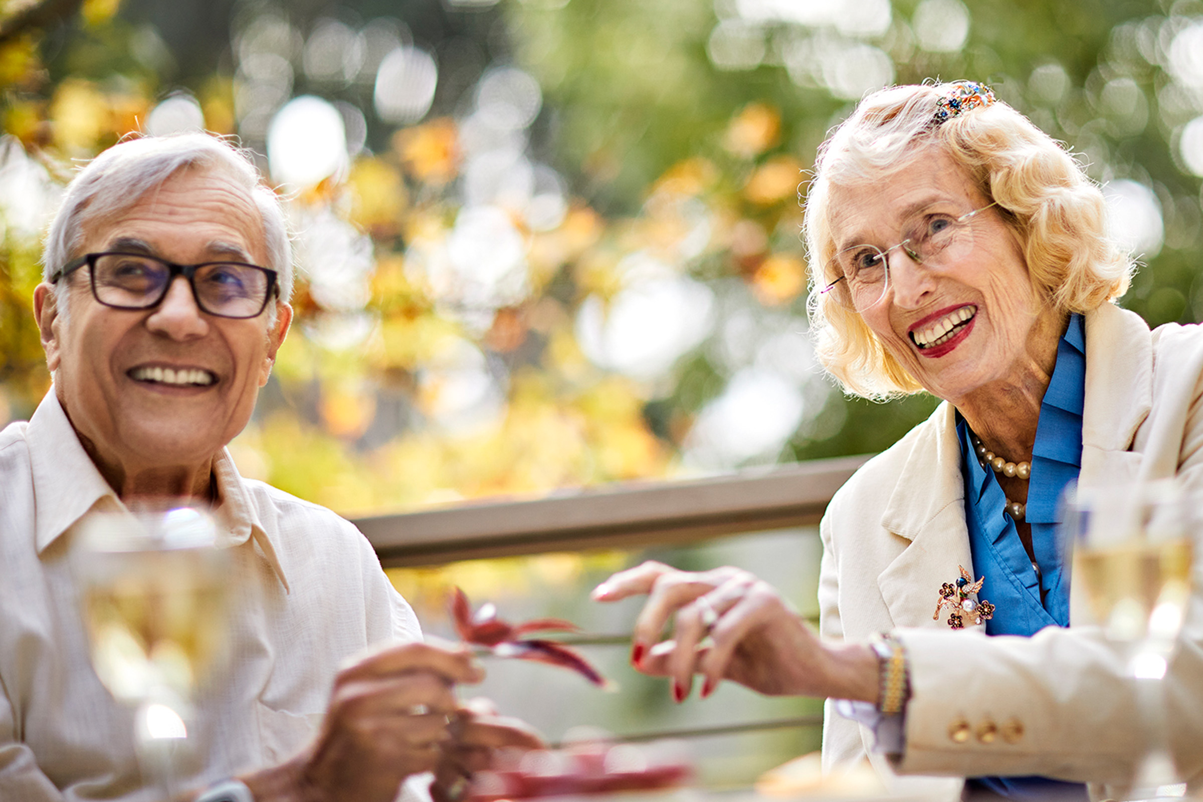 Two smiling older adults sit outdoors at a table, enjoying each others company. Both wear glasses and light-colored clothing, with wine glasses and plates in front of them. Sunlight filters through green and yellow leaves in the background.