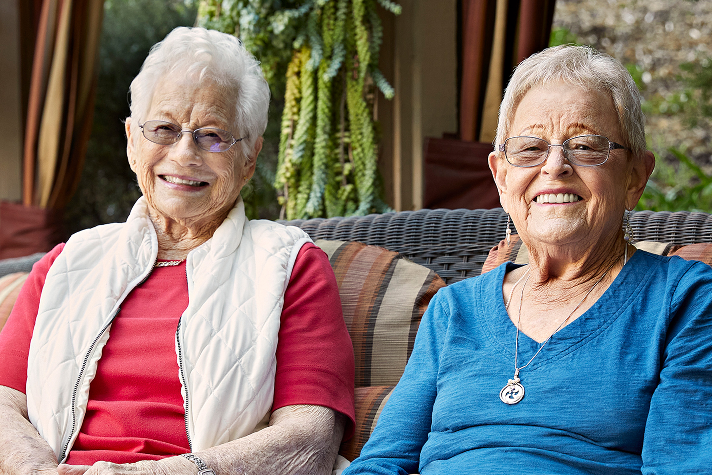 Two elderly women with short gray hair and glasses are smiling while sitting side by side on an outdoor wicker bench. One wears a red top with a white vest, the other a blue top. Green plants and curtains are in the background.