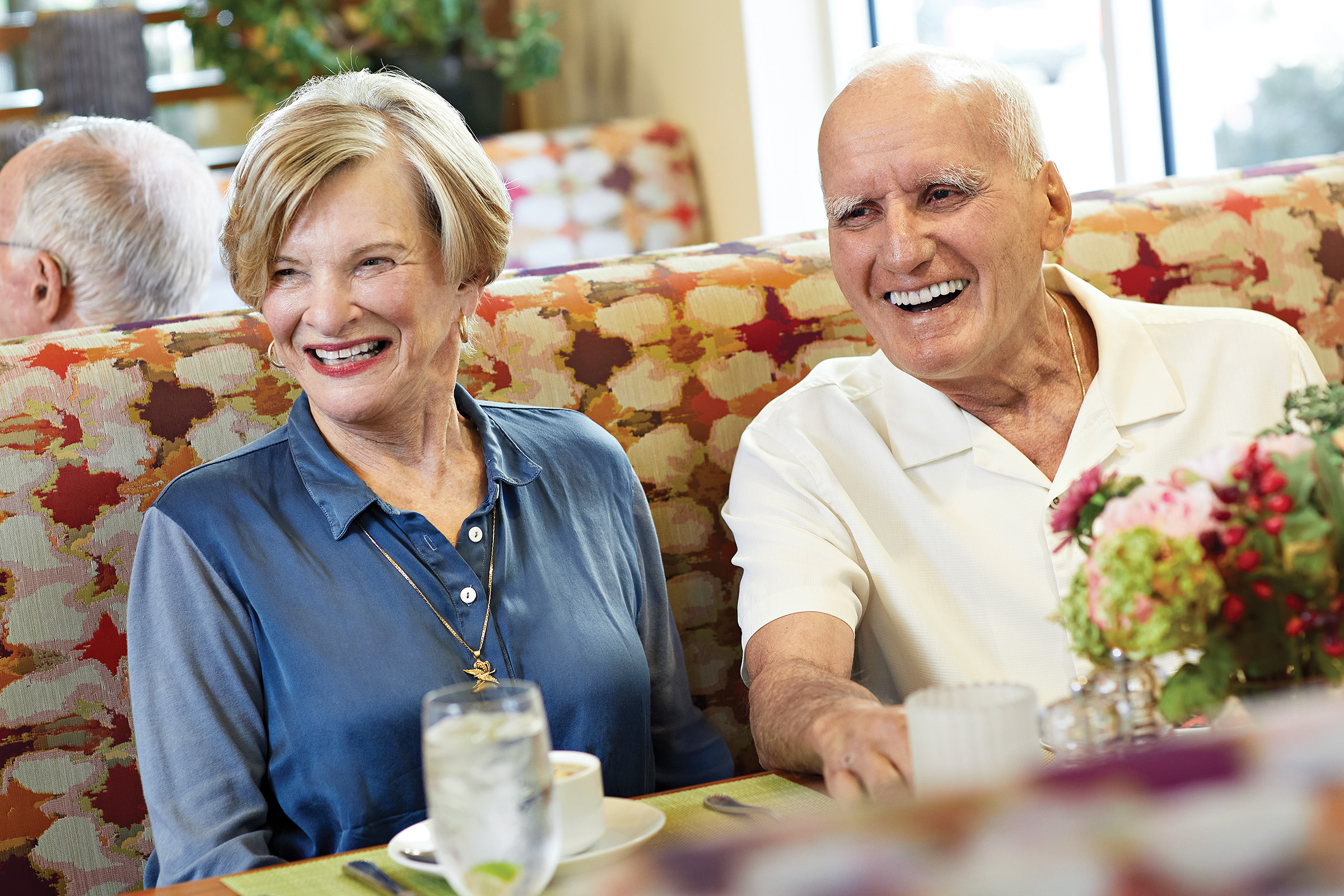 An older woman and man sit together in a brightly lit restaurant booth, smiling and laughing. A glass of water, flowers, and a cup are on the table in front of them.