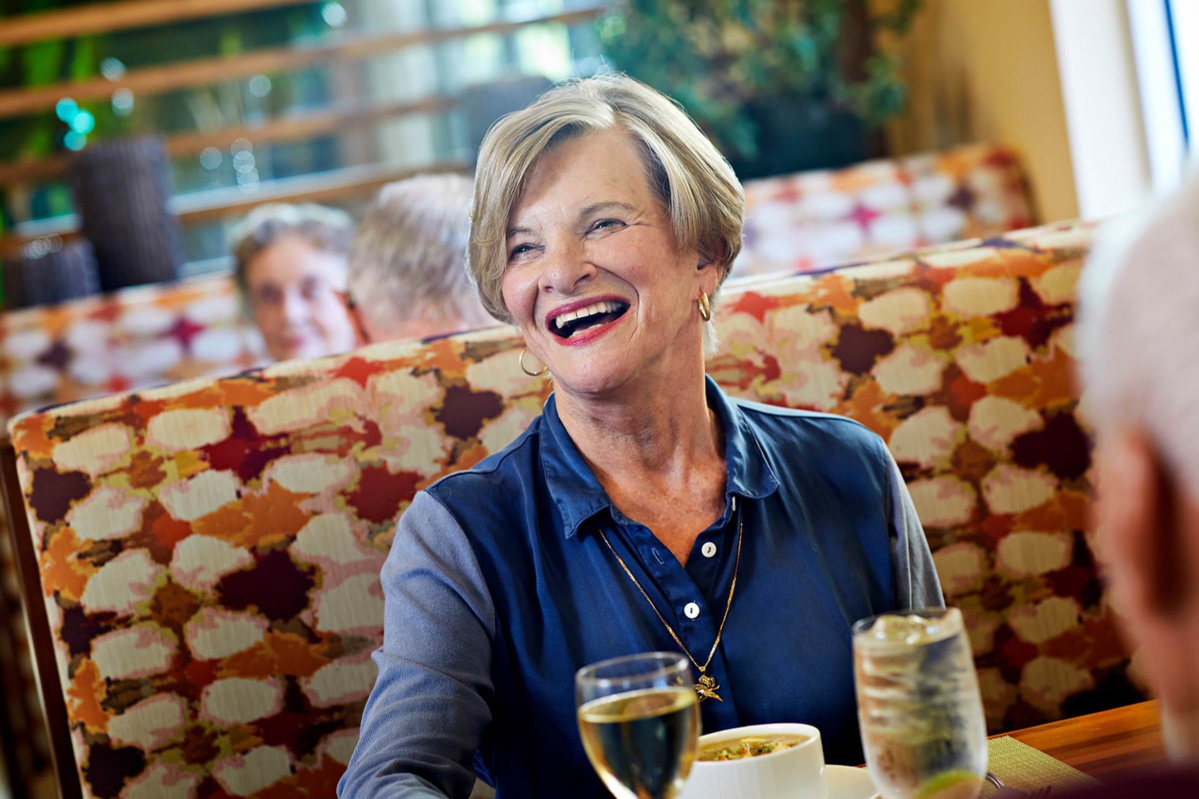 An older woman with short hair smiles while sitting at a restaurant table, enjoying a meal and drinks. There are other people in the background and colorful, patterned booth seating behind her.