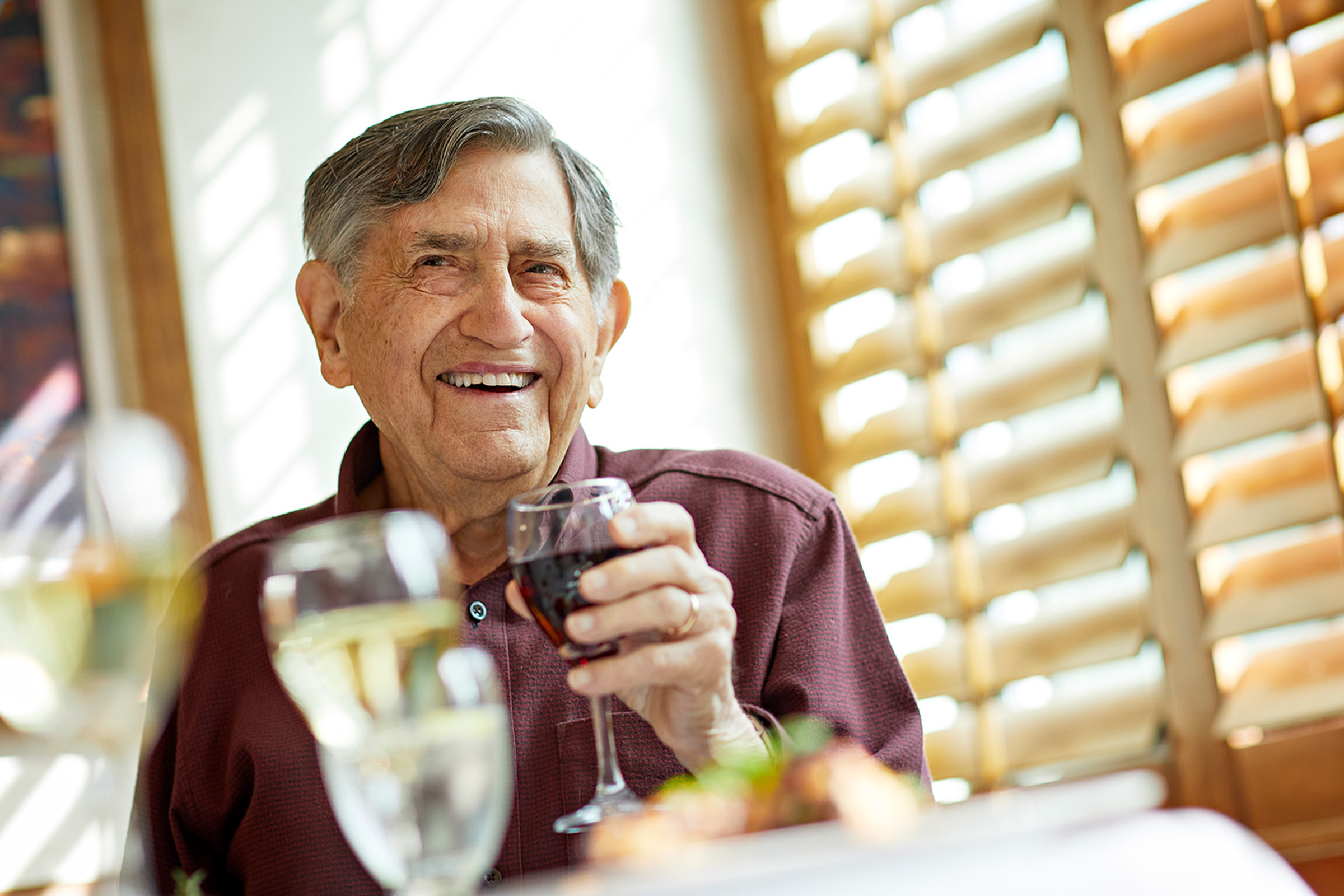 An elderly man with gray hair smiles warmly while holding a glass of red wine at a table set with drinks and food, seated in a sunlit room with wooden blinds.