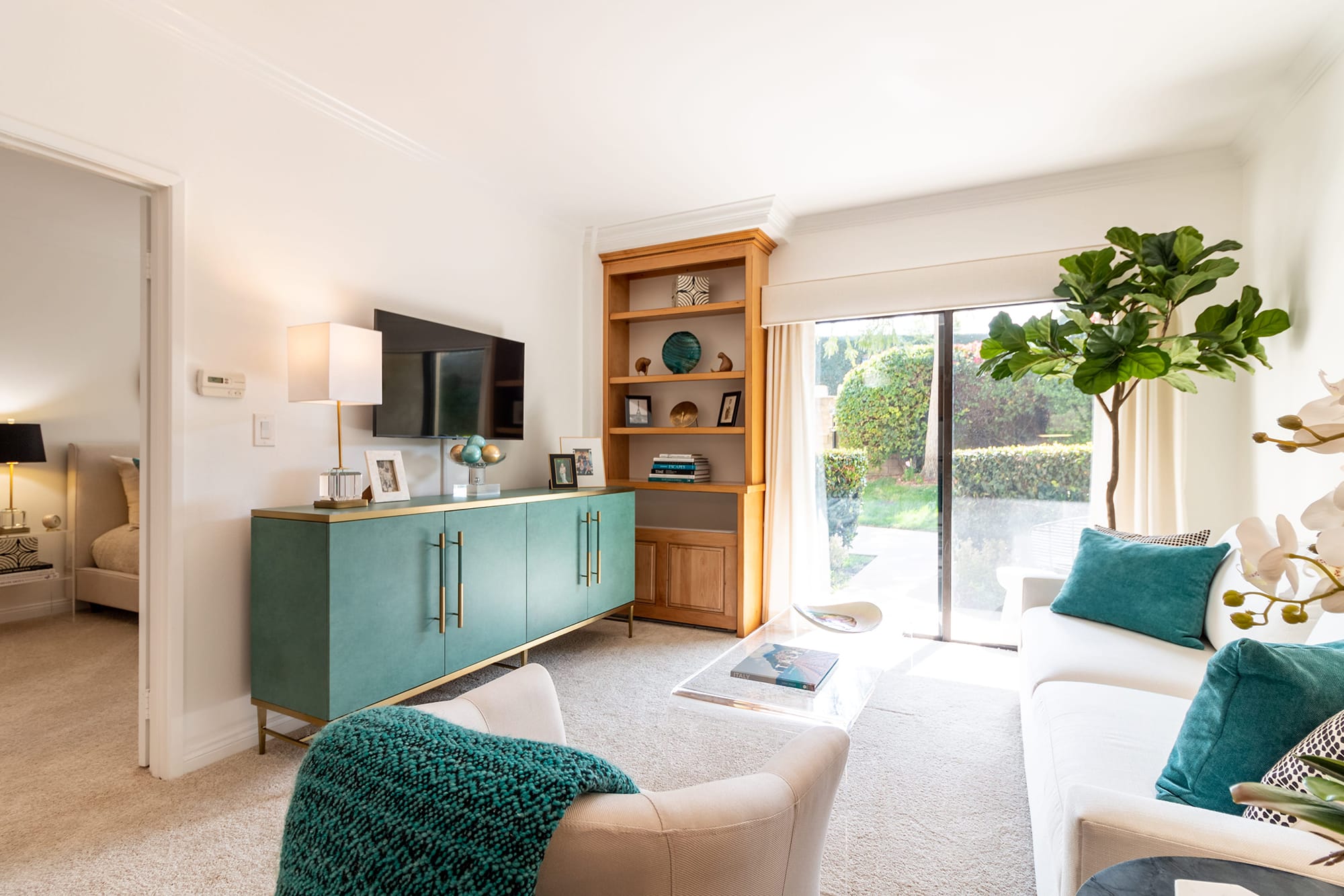 A bright living room with a white sofa, teal pillows, a teal sideboard, wall-mounted TV, wooden shelves, large potted plant, and sliding glass doors opening to a green garden. A cozy bedroom is visible through an open door.