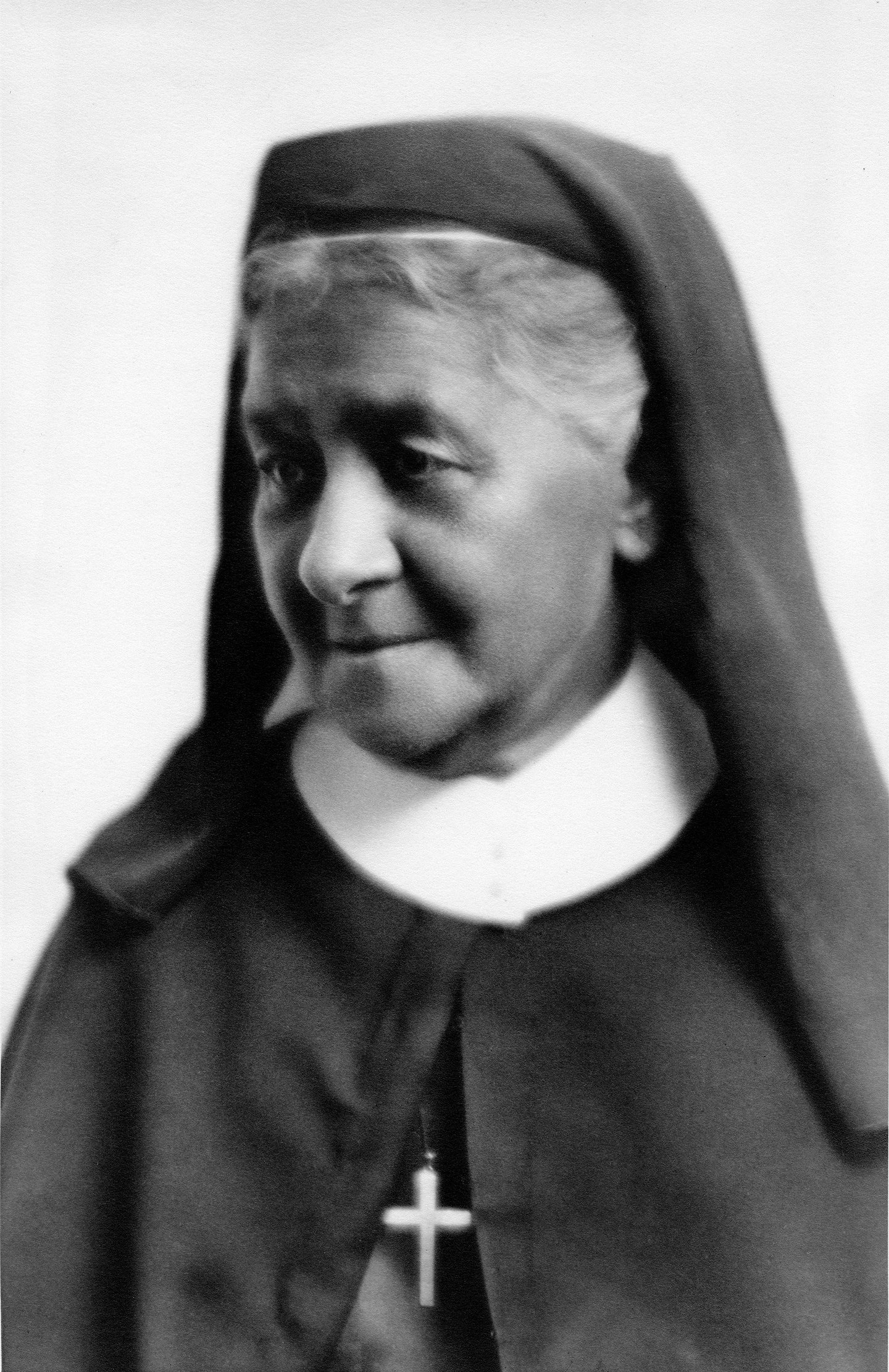 Black and white portrait of an elderly woman in a nun’s habit, wearing a dark veil, white collar, and a cross necklace, looking slightly to the side with a gentle expression.