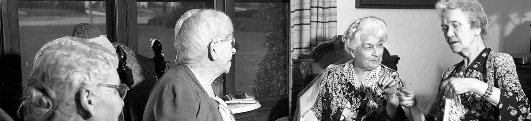 Four elderly women sit together in a room, engaged in conversation. Two women face each other while the others sit nearby, all in vintage clothing. Large windows and framed pictures are visible in the background.