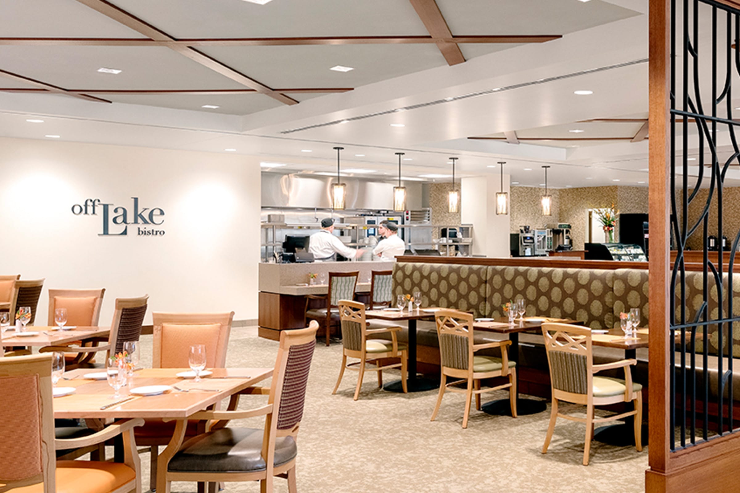 A modern restaurant dining area with wooden tables and chairs, upholstered booth seating, and an open kitchen where two chefs are working under bright lights. The wall displays the sign off Lake bistro.