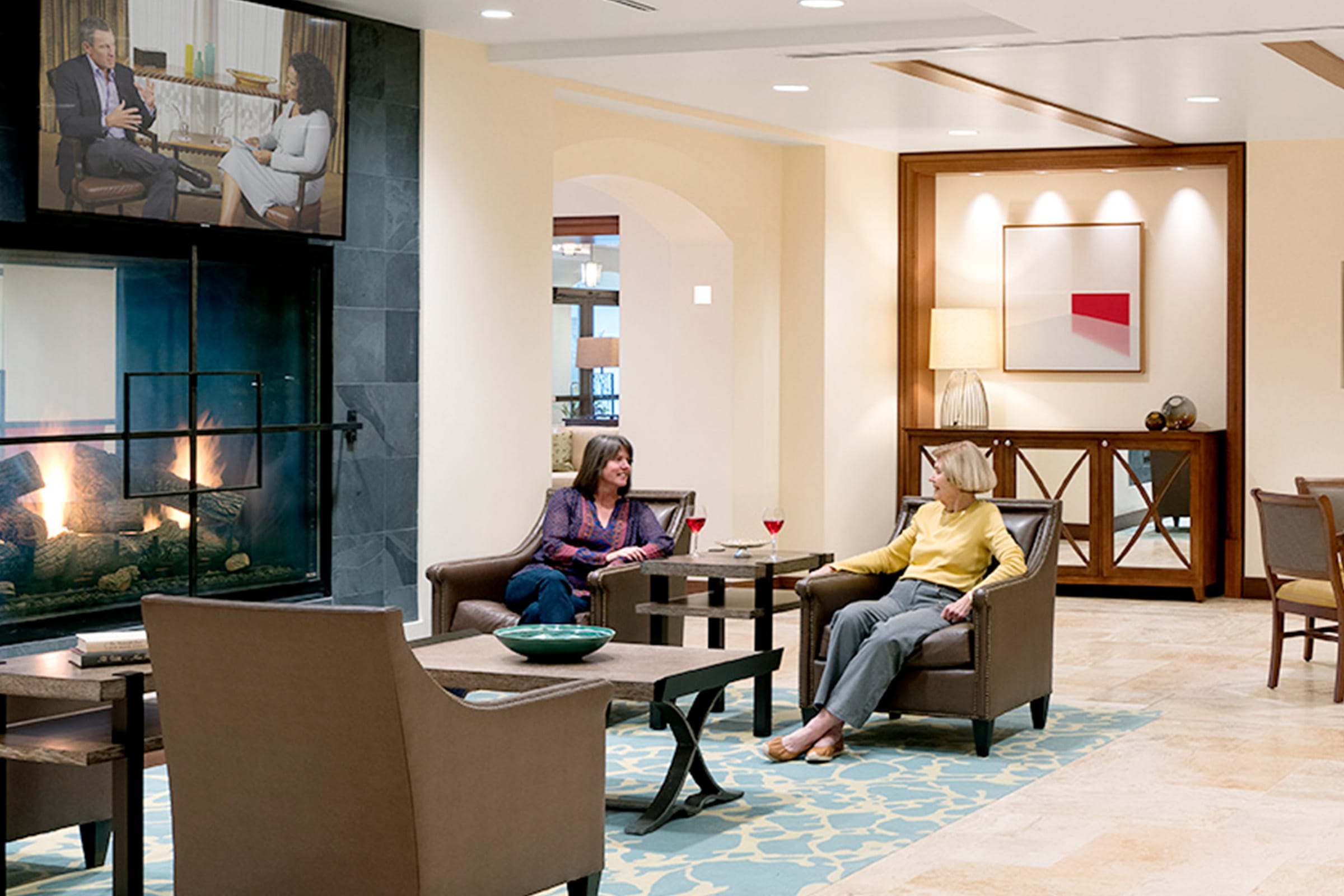 Two women sit and talk in a modern lounge with a fireplace. A TV above the fireplace displays a talk show. The room has armchairs, a rug, art on the wall, and warm lighting.