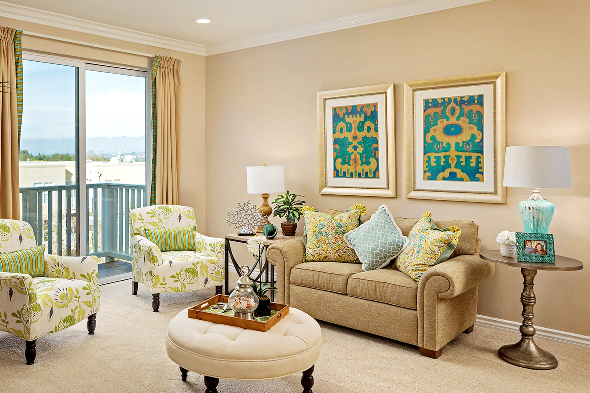 A cozy living room with a beige sofa, patterned pillows, two floral armchairs, a round ottoman, and a small table lamp. Two colorful abstract artworks hang on the wall above the sofa. Glass doors open to a balcony.
