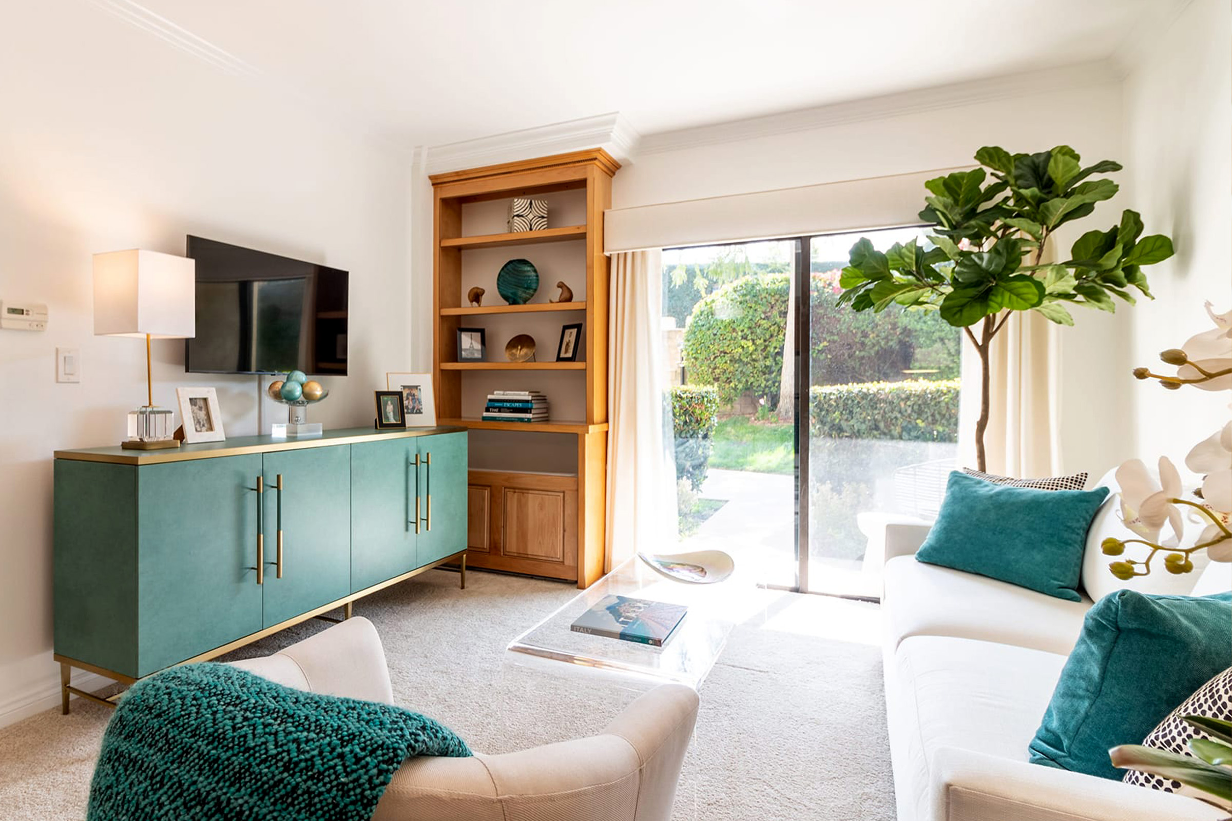 Bright living room with a white sofa, teal cushions, green sideboard, wall-mounted TV, wooden shelving, large plant, glass coffee table, and sliding glass doors opening to a garden.