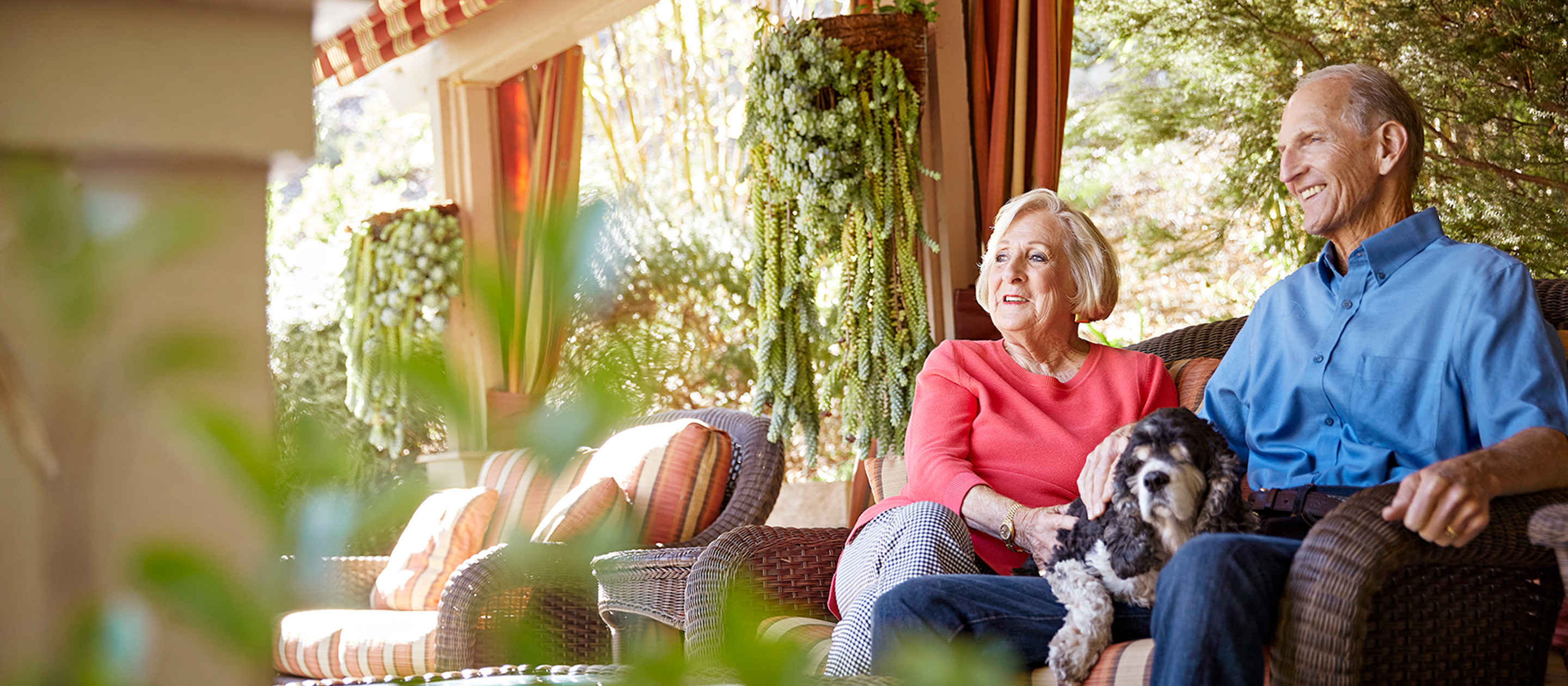 An elderly couple sits on a wicker sofa outdoors, smiling and relaxing together. The woman, wearing a red top, holds a small black and white dog on her lap. Lush greenery and hanging plants decorate the background.