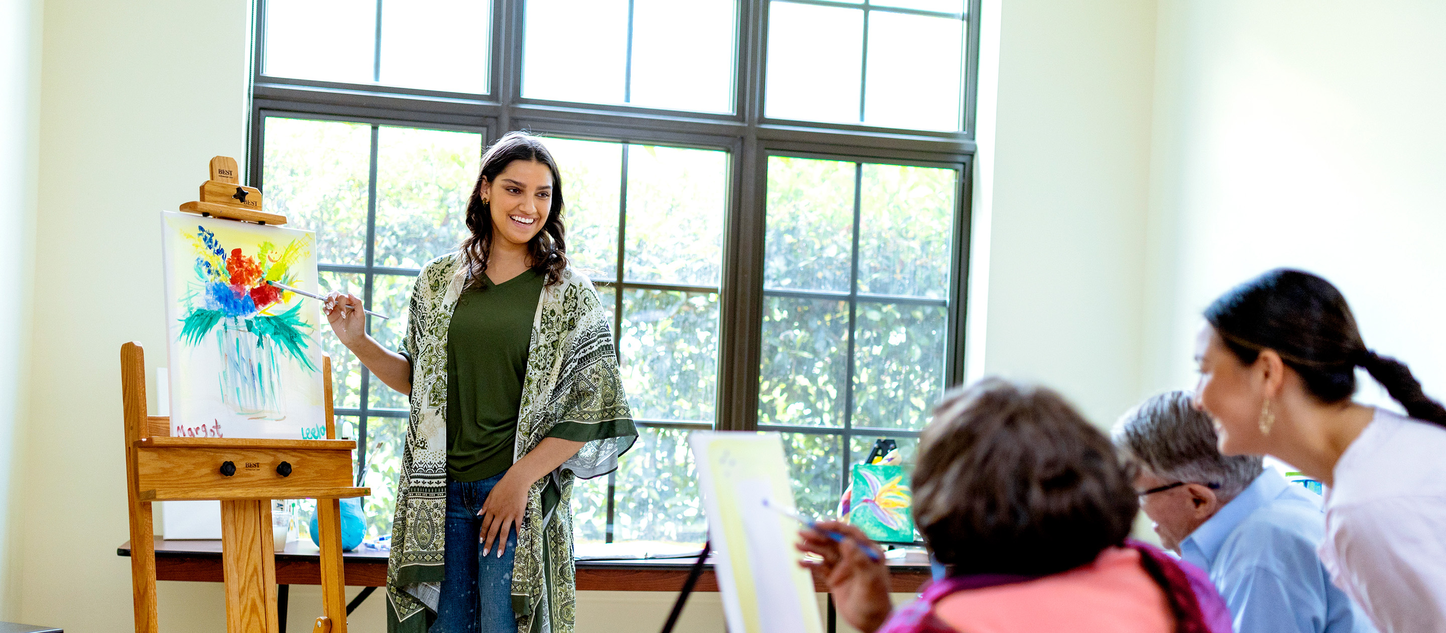 A woman stands at an easel, painting a colorful floral scene, while smiling at a group of seated people who are watching and painting along in a bright room with large windows.