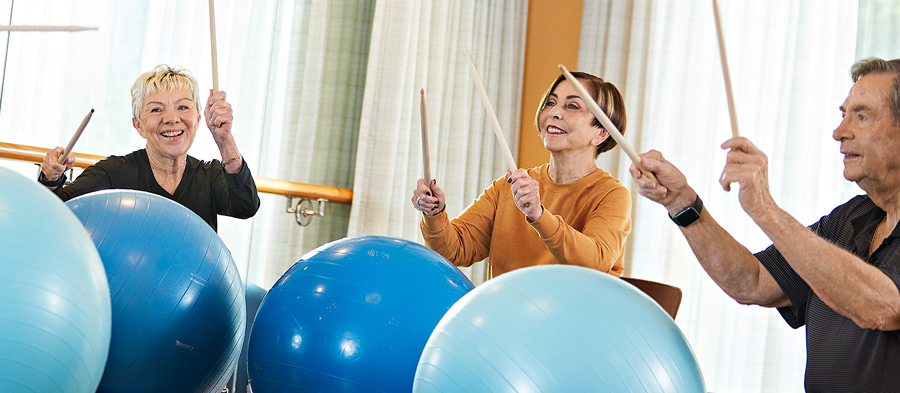 Three older adults participate in a fitness class, smiling as they use drumsticks to tap large blue exercise balls in a bright, sunlit room with curtains in the background.