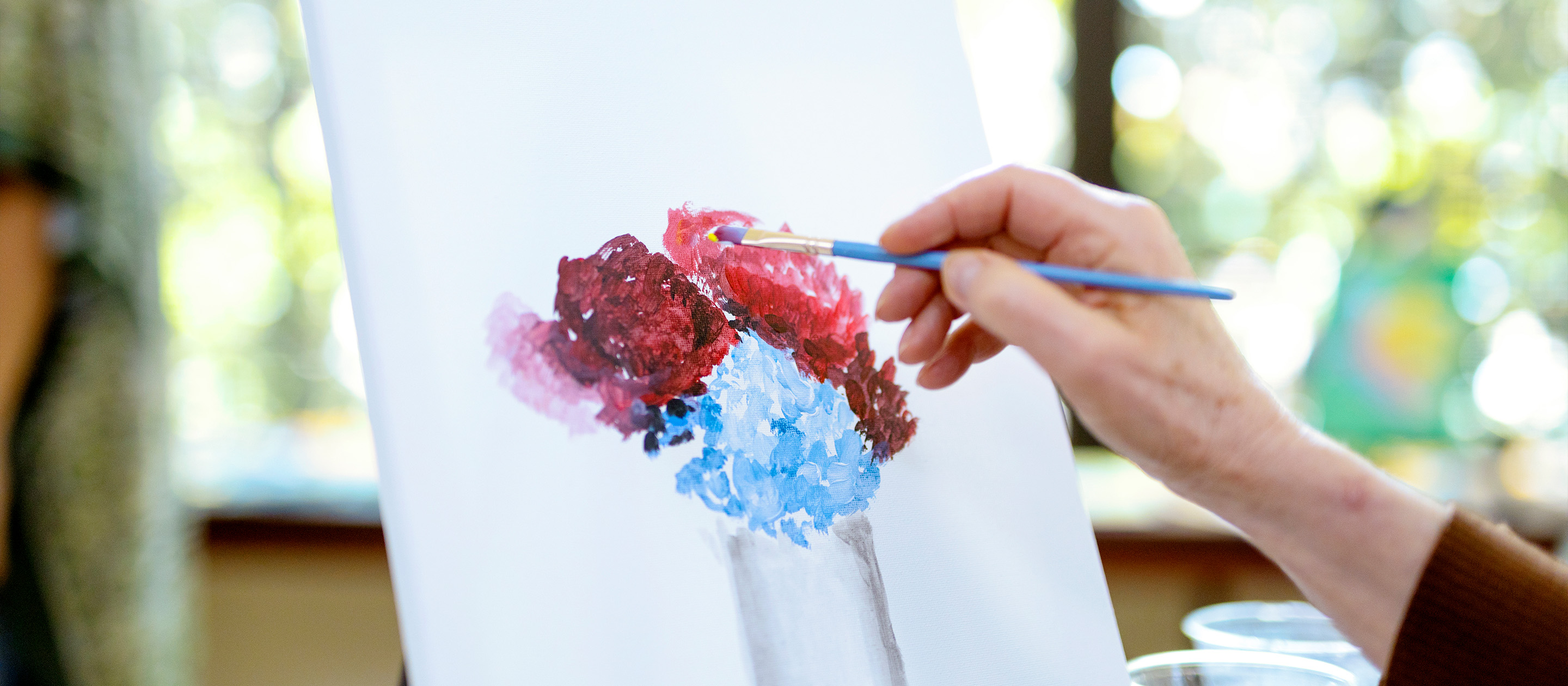 A person’s hand holds a paintbrush, painting a bouquet of red and blue flowers on a white canvas. The background is blurred, showing soft natural light and greenery through a window.