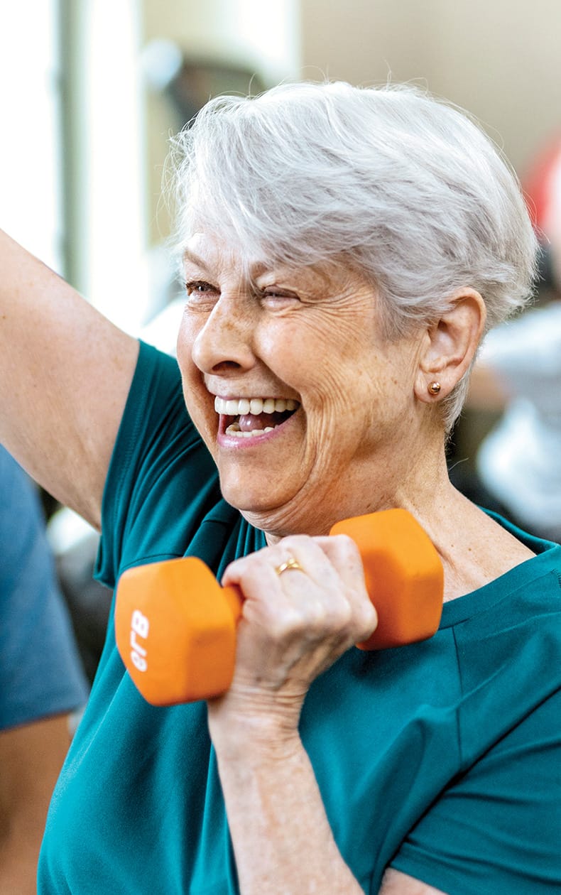 Smiling older woman with short gray hair lifts an orange dumbbell while exercising, wearing a teal shirt, in a bright indoor setting.
