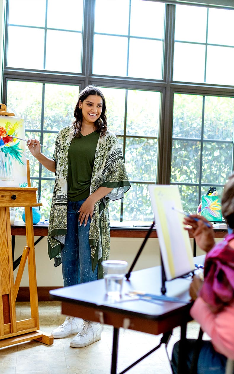 A woman stands by an easel with a colorful painting, smiling and teaching an art class. Another person sits at a table painting. Large windows and natural light fill the room.