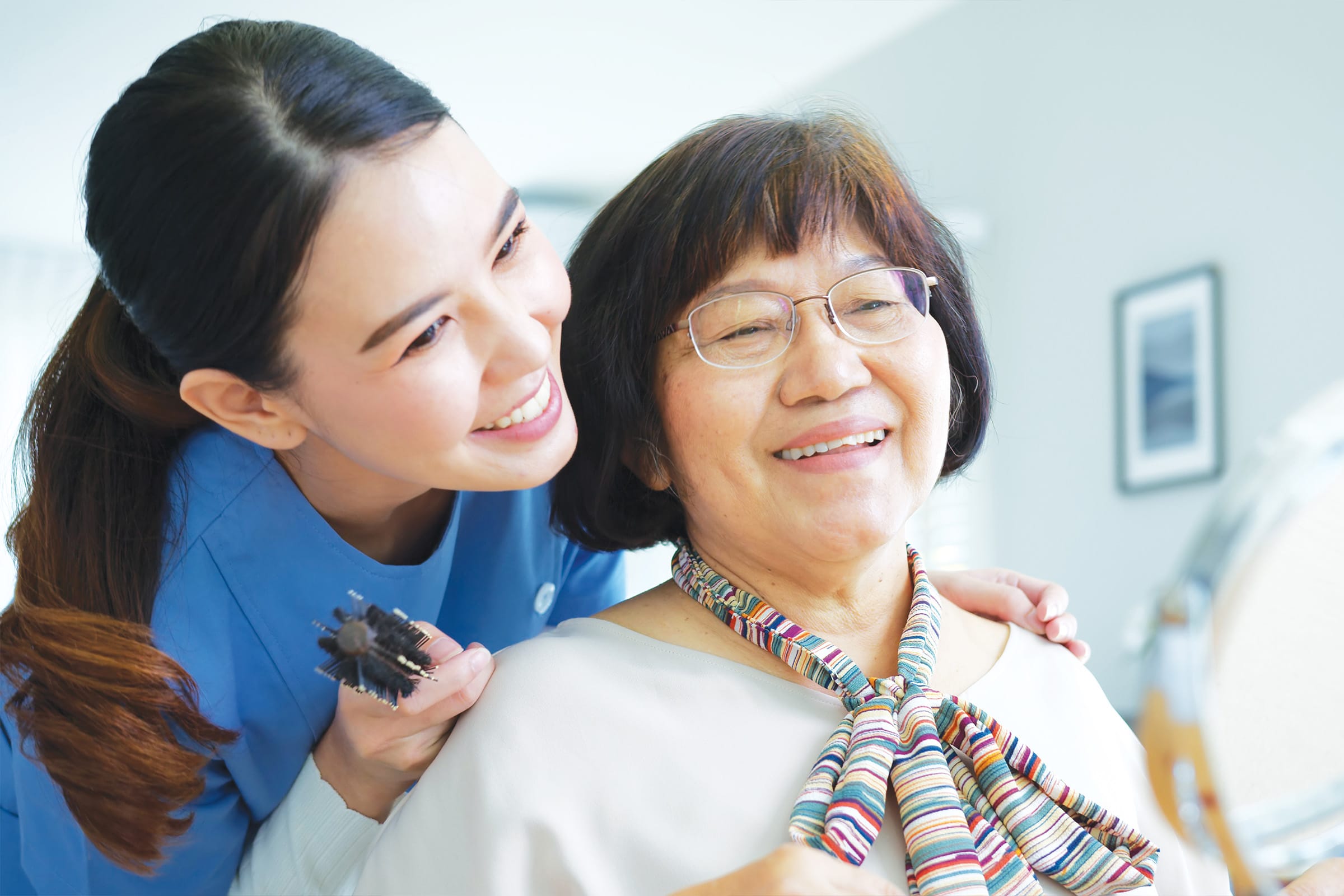 A smiling caregiver in blue scrubs holds a hairbrush while standing beside an older woman with glasses and a colorful scarf, who is also smiling and looking into a mirror.