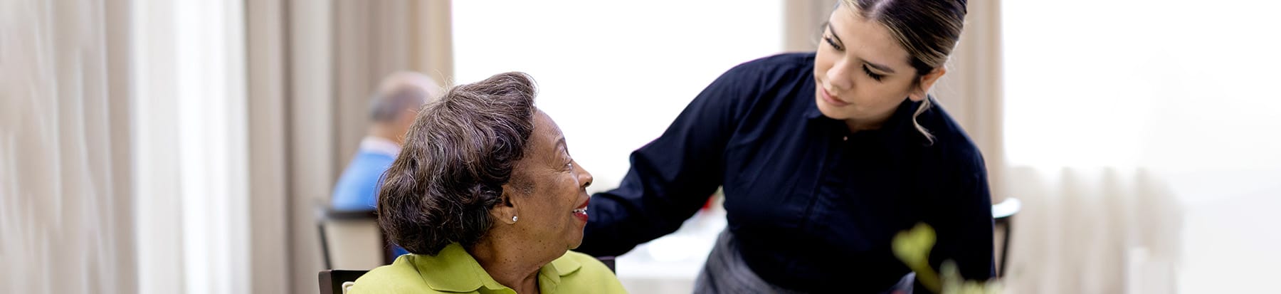 A young woman in a dark uniform leans down to talk with an older woman in a green jacket who is seated at a table, both smiling warmly in a bright, softly lit room.