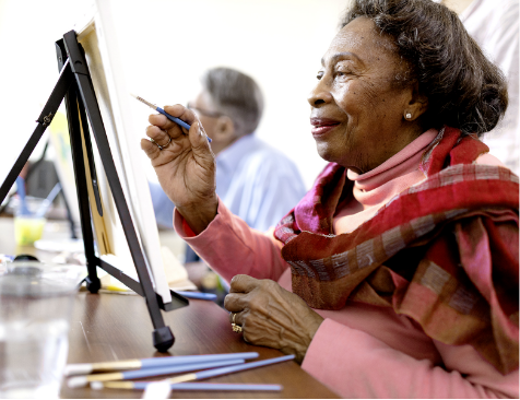 An elderly woman smiles while painting on a canvas at an easel, wearing a pink sweater and scarf. Colored pencils are on the table, and another person is painting in the background.