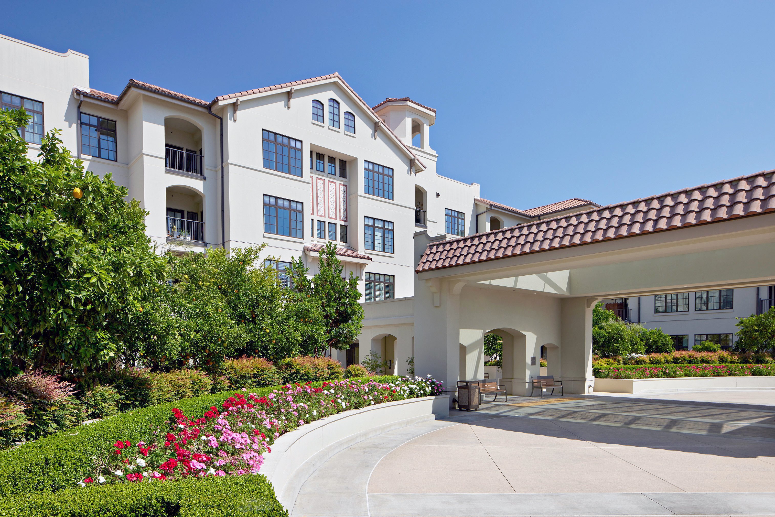 A large, modern apartment or hotel building with white walls and red-tiled roofs, surrounded by lush landscaping, colorful flowers, and a covered driveway under a clear blue sky.