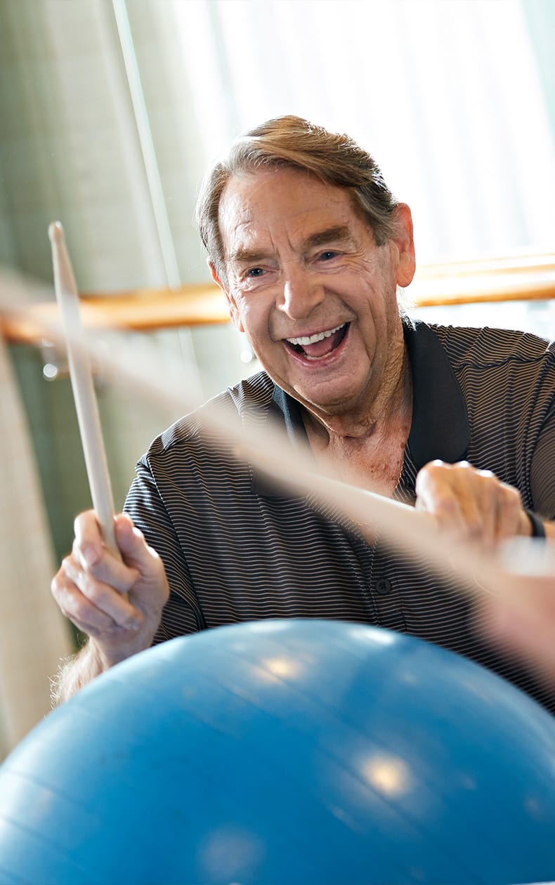 An older man smiles while rhythmically hitting a large blue exercise ball with drumsticks, appearing energetic and joyful during an indoor fitness activity.