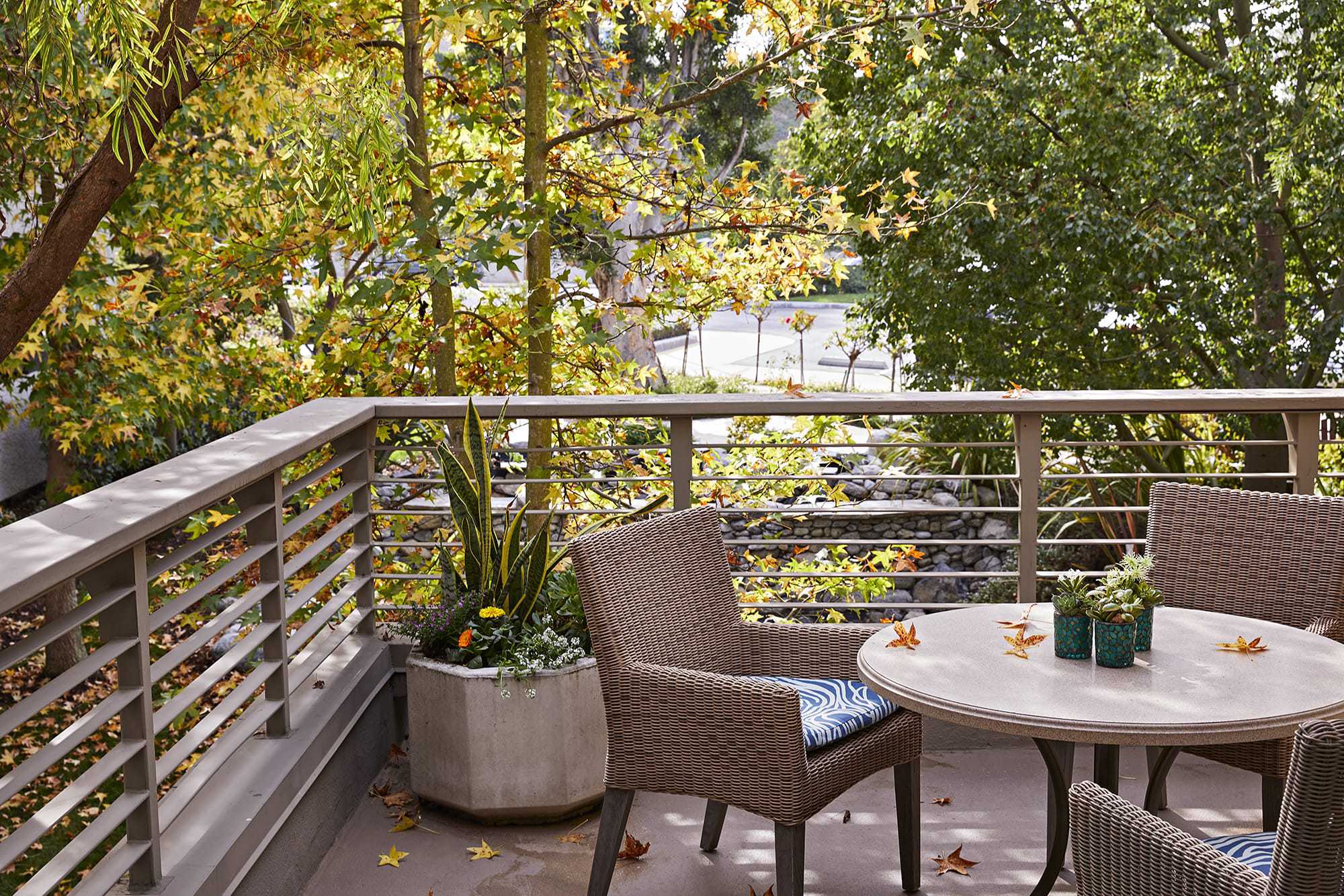 Outdoor patio with a round table and woven chairs, some autumn leaves on the table and floor. Green and yellow trees surround the area, creating a peaceful, leafy atmosphere. A large planter sits in the corner.