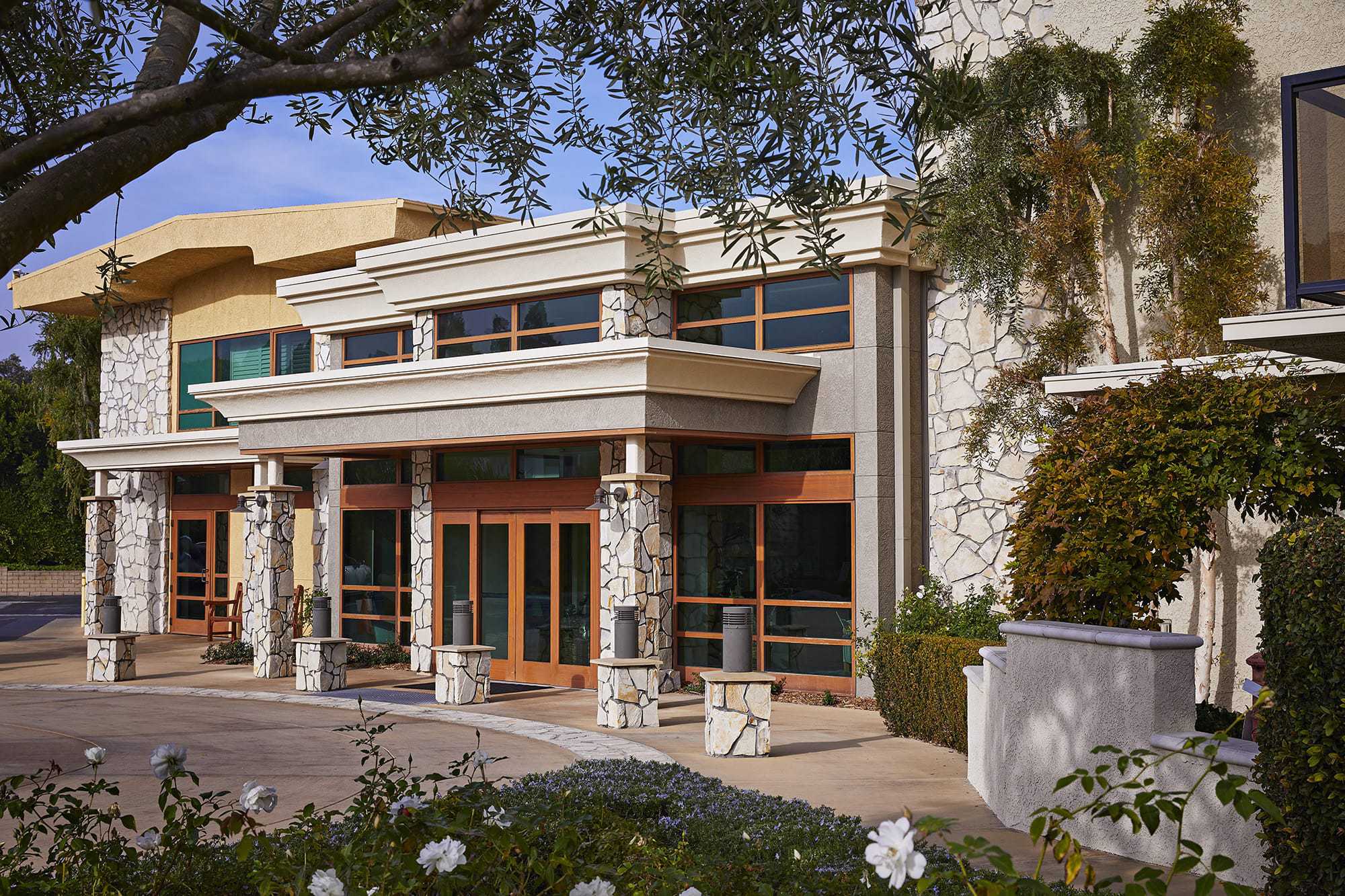 Modern building entrance with large wooden-framed glass doors, stone pillars, and beige exterior walls. Shrubs, flowers, and trees decorate the foreground, and sunlight casts shadows across the scene.