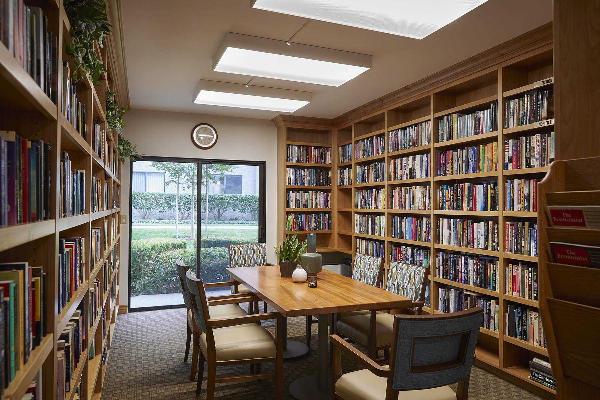 A cozy library room with bookshelves lining the walls, a wooden table with six chairs in the center, potted plants on the table and shelf, large window with a view of greenery, and a clock above the window.