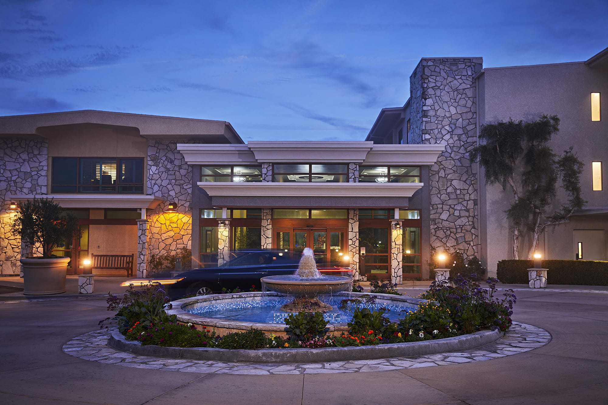 A round fountain with flowers in front of a modern building with stone accents, large windows, and exterior lights at dusk. A dark car is parked near the entrance.