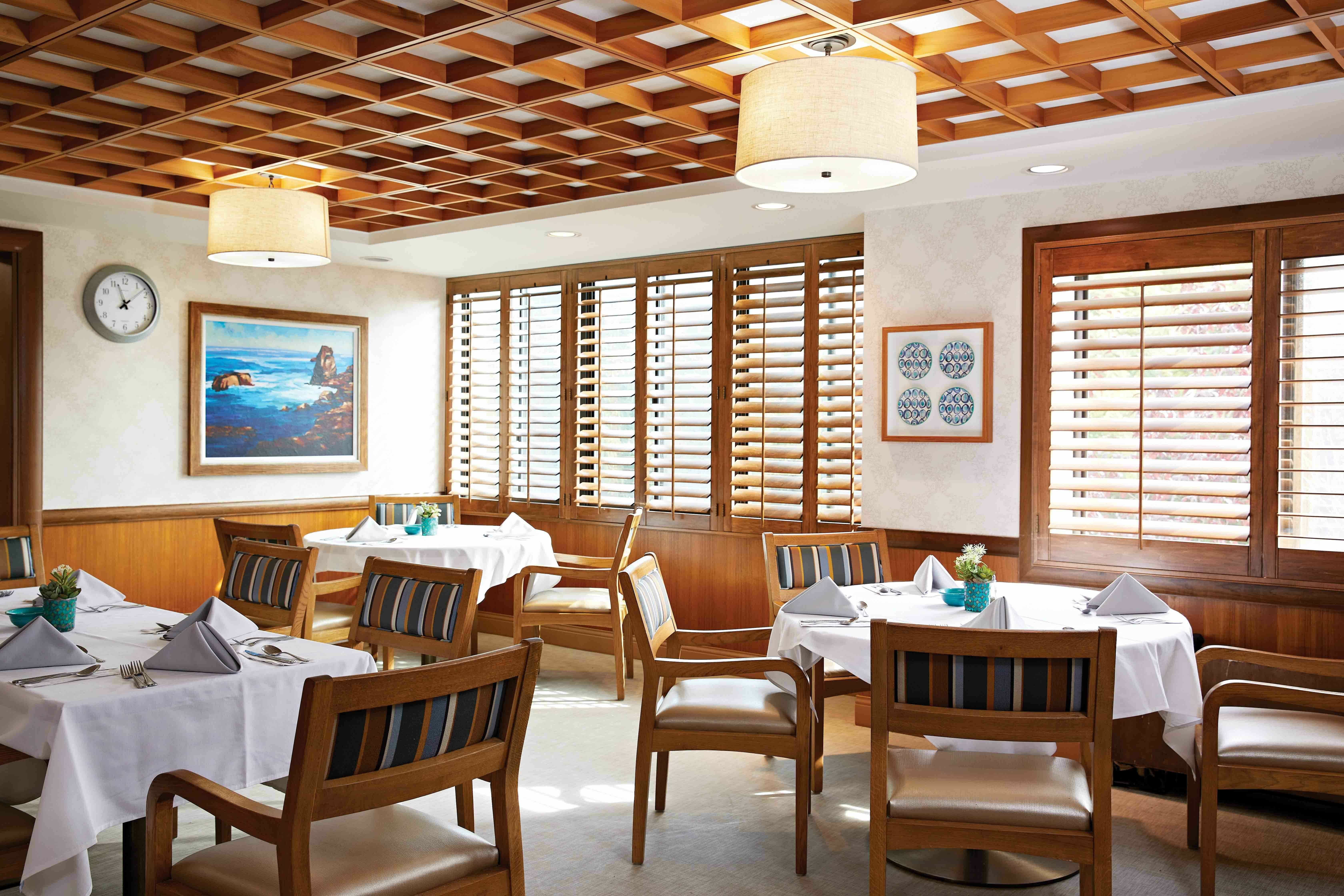 A bright, cozy restaurant dining area with wooden chairs around tables covered in white cloths, napkins folded on each table, large windows with wooden shutters, a clock, and artwork on the walls.