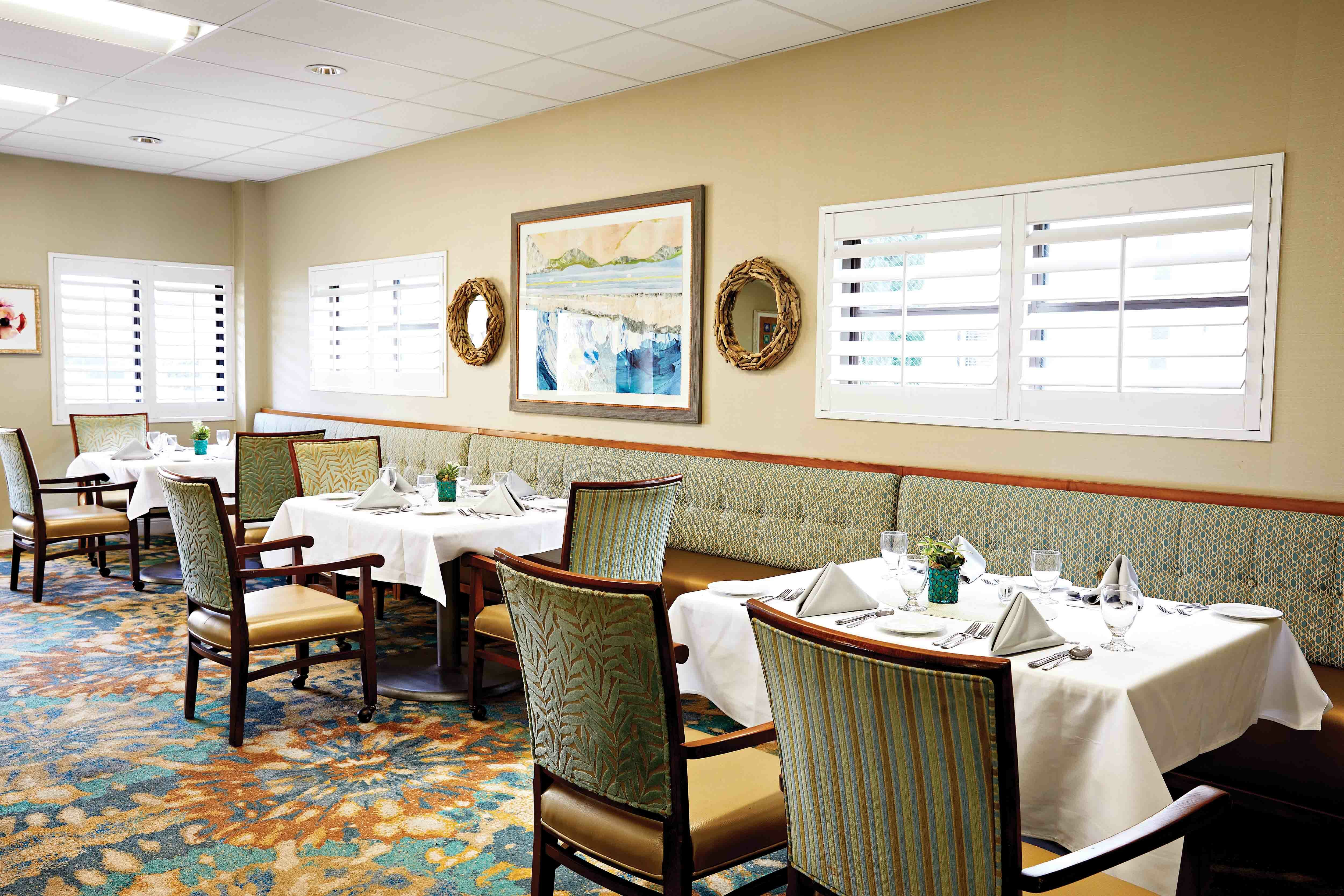 A bright, elegant restaurant dining area with tables set with white cloths, napkins, and glassware. Upholstered chairs and a long bench line the wall, which features two windows and framed artwork.