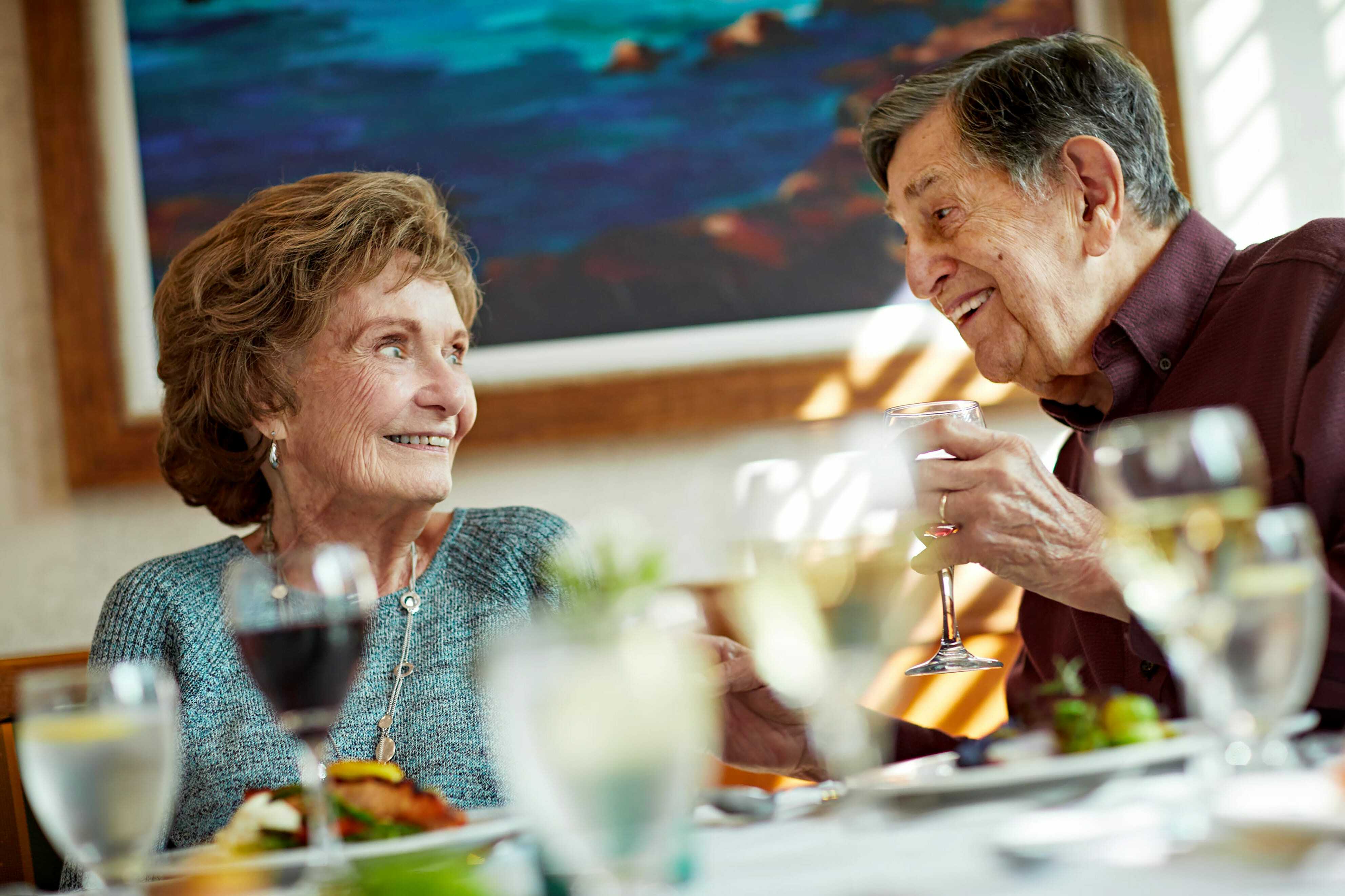 An elderly couple sits at a table, smiling and enjoying a meal together in a warmly lit restaurant. Glasses of wine and plates of food are visible in the foreground.