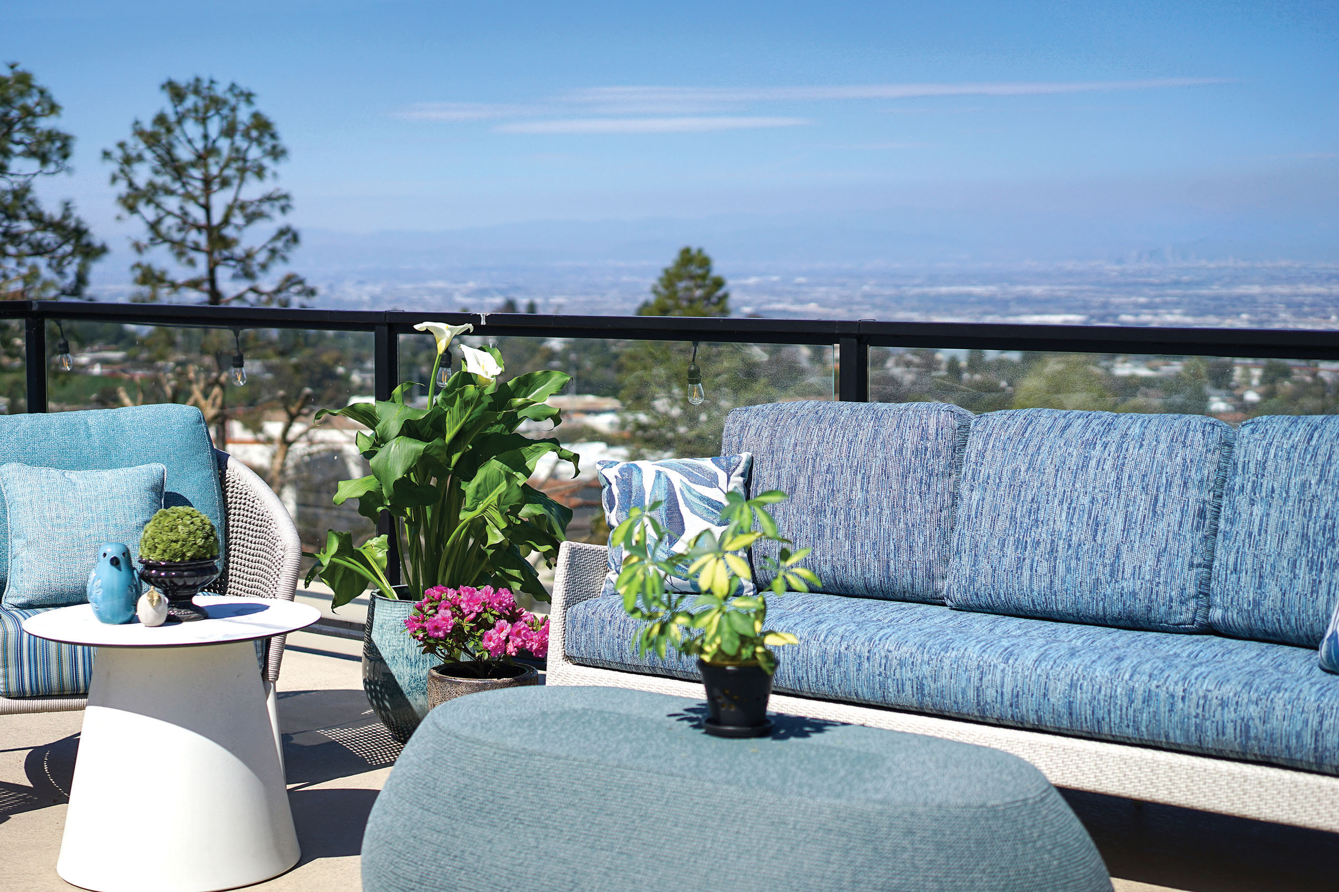 Outdoor patio with blue and white furniture, a small white table with a bird figurine, potted plants, and flowers. Glass railing overlooks a scenic city view with trees and a clear, sunny sky.