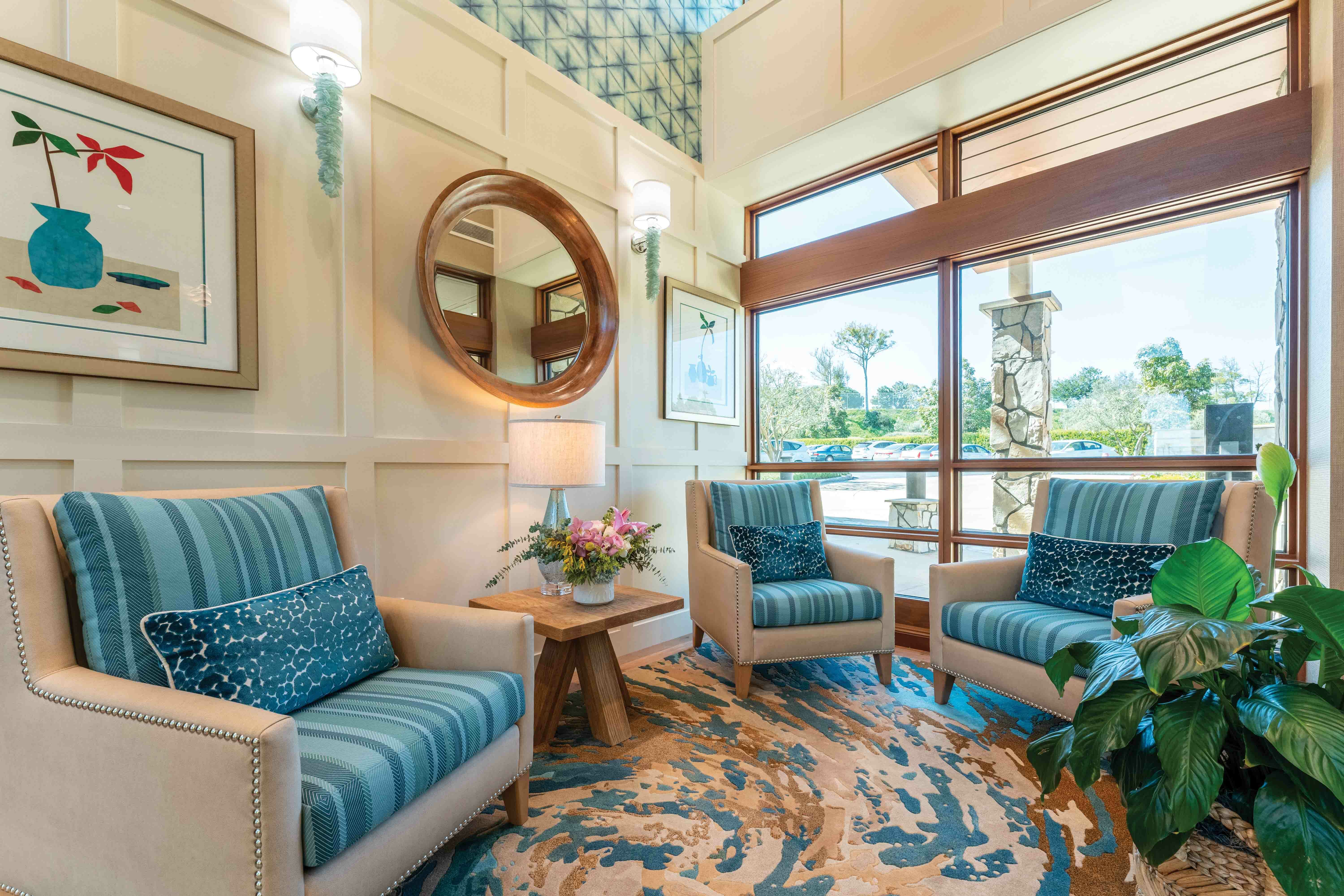 A cozy seating area with three blue armchairs, a wooden side table with a flower arrangement, large windows letting in natural light, wall art, and a round mirror above the chairs. Indoor plants decorate the scene.