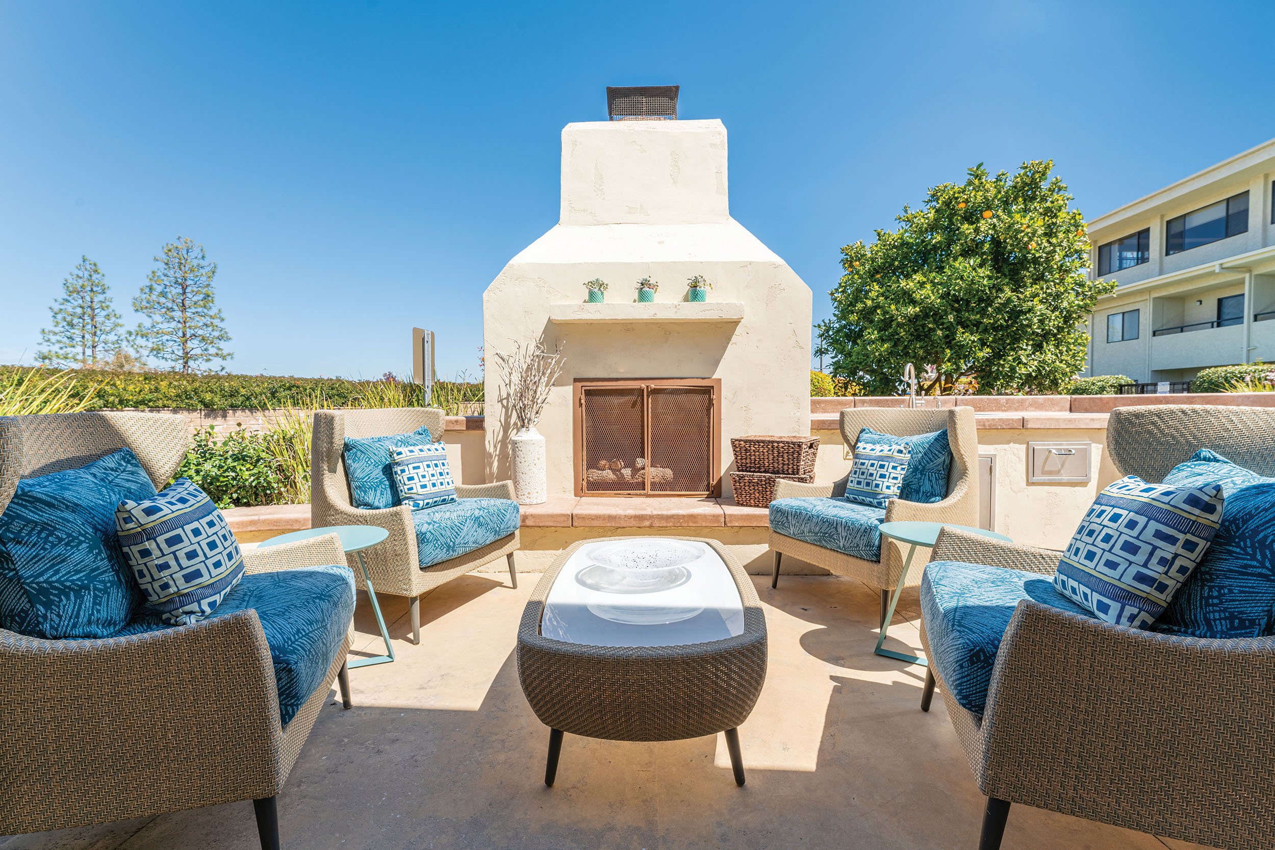 Outdoor patio with cushioned chairs arranged in a circle around a small table, facing a white stucco fireplace. Pillows decorate the chairs, and trees and a building are visible in the background under a clear blue sky.