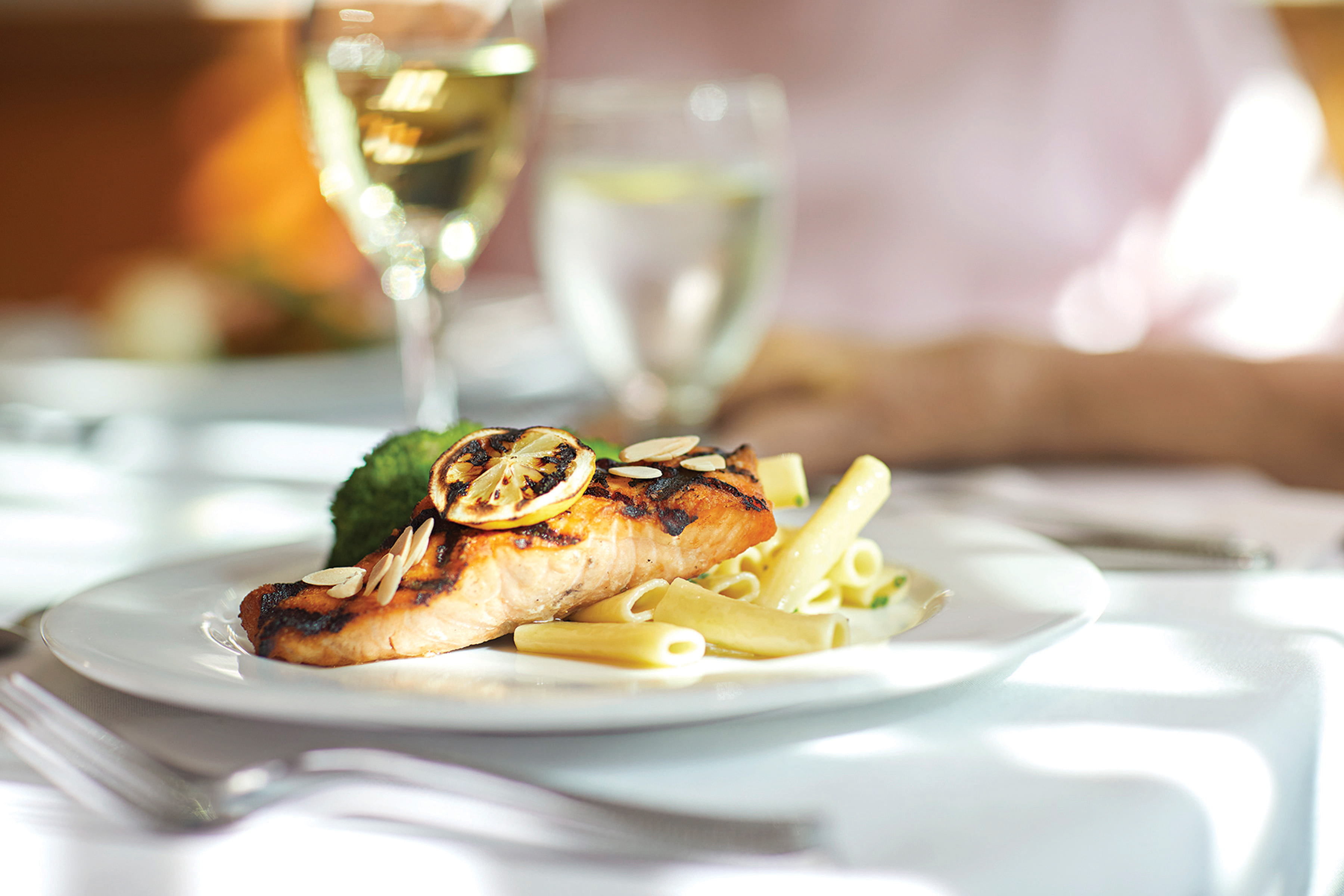 A close-up of a plate with grilled salmon topped with lemon slices and almonds, served with penne pasta. A glass of white wine and a glass of water are in the background on a white tablecloth.