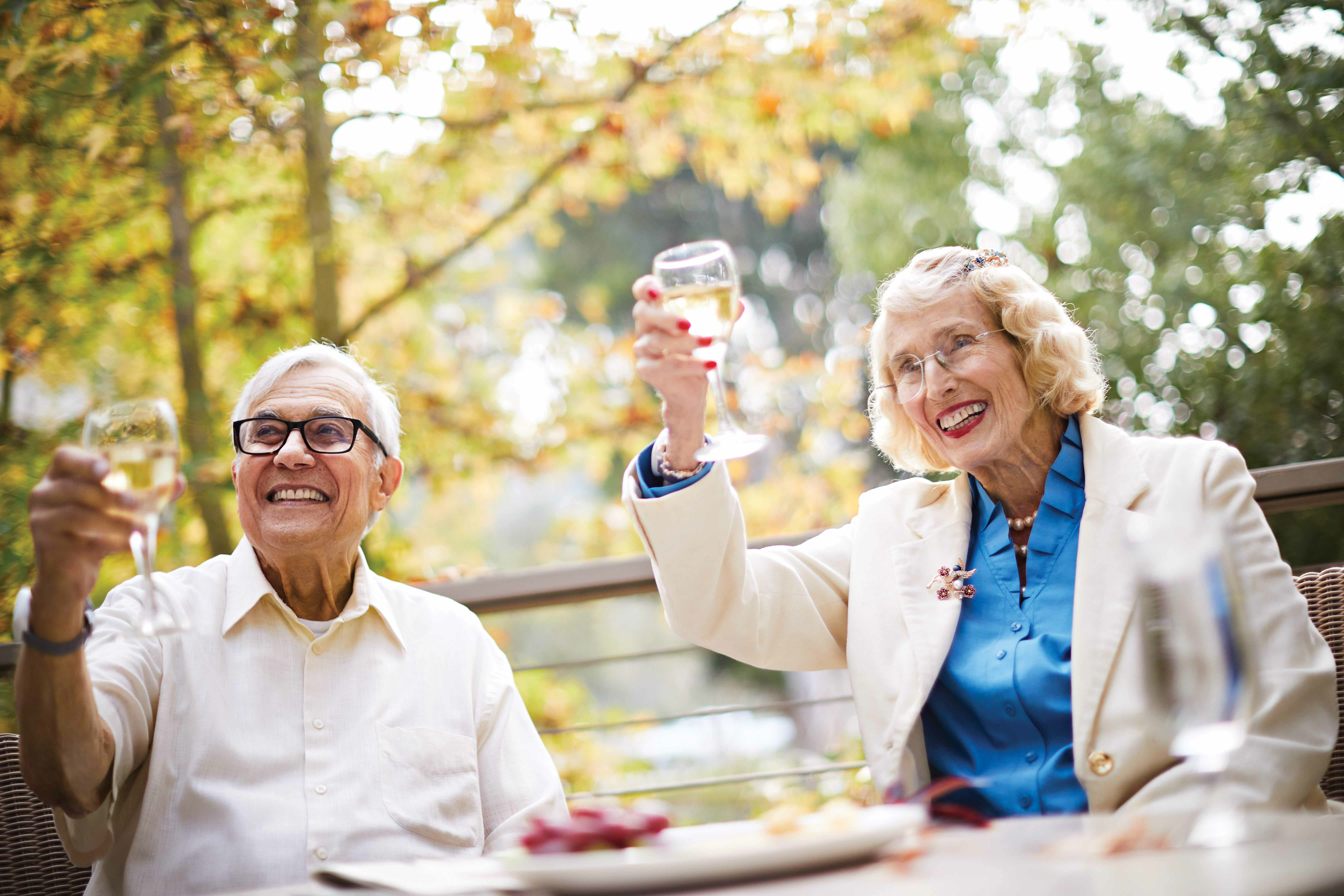 Two smiling older adults, a man and a woman, sit outdoors at a table, raising glasses of white wine in a toast. They are surrounded by trees with yellow leaves, suggesting a pleasant autumn day.