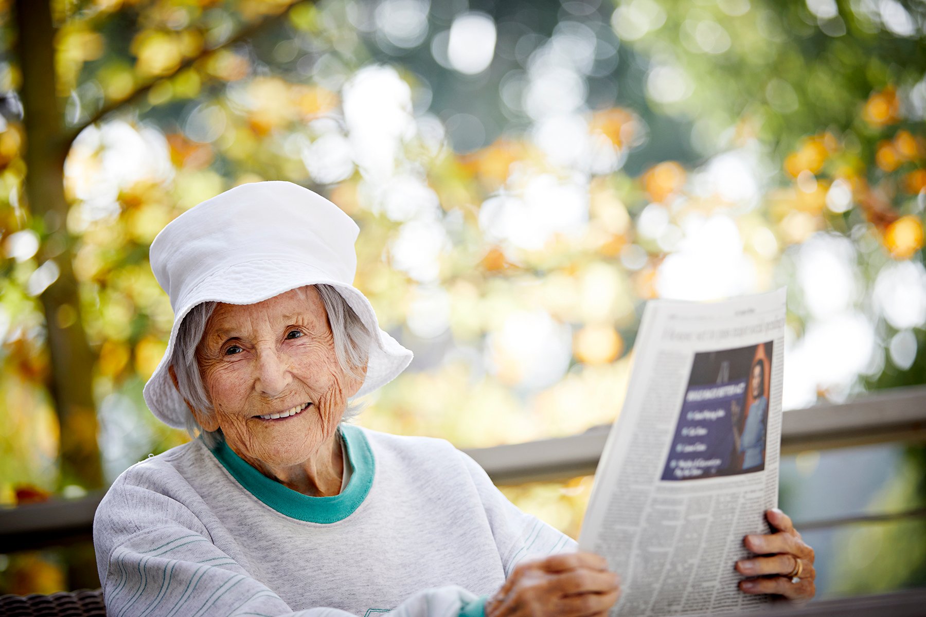 An elderly woman wearing a white hat and light sweater sits outdoors, smiling and holding a newspaper. Sunlight filters through trees in the background, creating a warm, cheerful atmosphere.