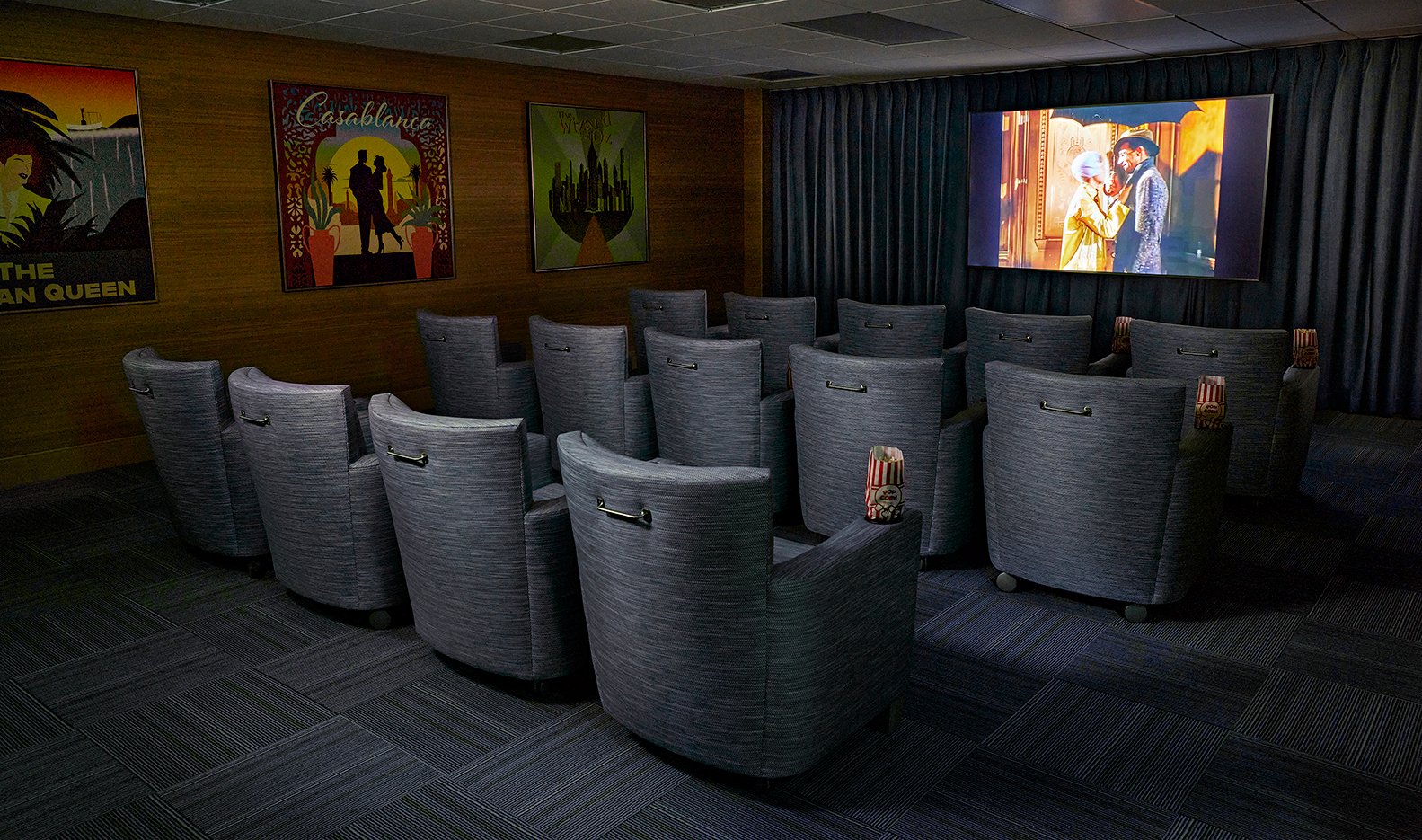 Small private movie theater with three rows of cushioned seats, each with popcorn, facing a screen showing a classic film. Movie posters decorate the wood-paneled walls, and the room is dimly lit.