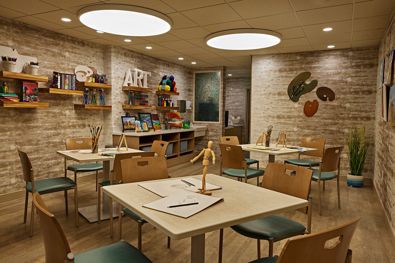 A well-lit art classroom with tables and chairs, art supplies on shelves, drawing mannequins, and sketchpads. The walls are decorated with the word ART and colorful palettes. A small plant is in the corner.
