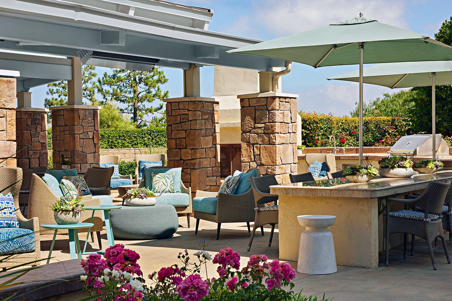 Outdoor patio with wicker chairs, blue and patterned cushions, large umbrellas, a stone bar, potted plants, and blooming pink flowers in the foreground. The area is bright and inviting under a sunny sky.