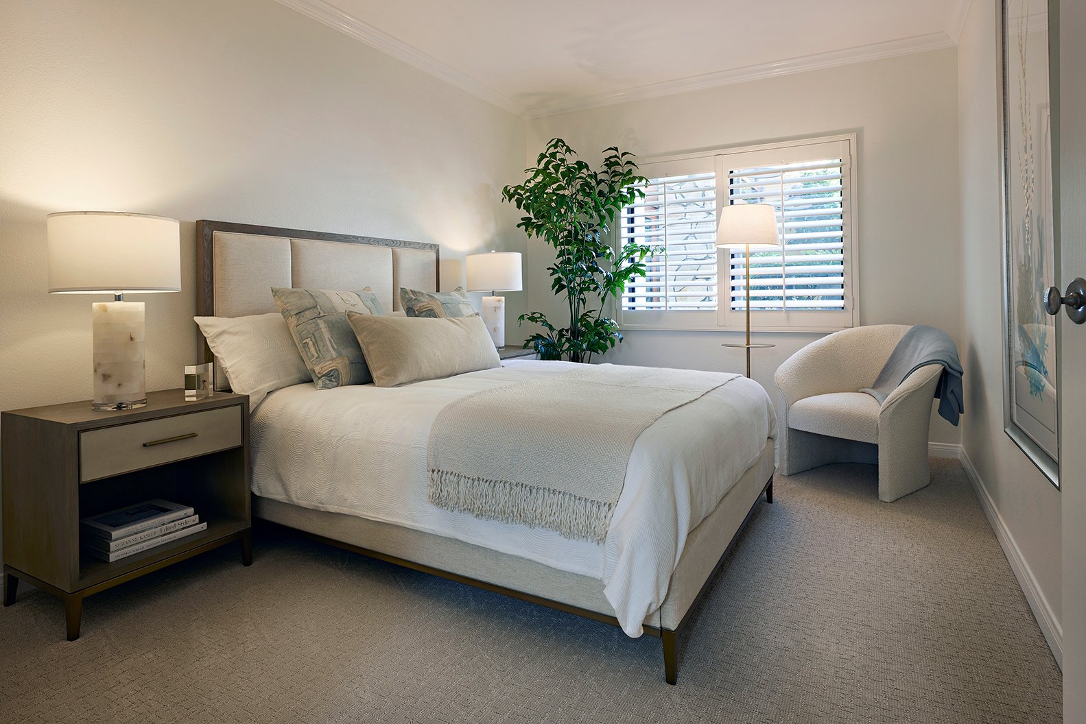 Modern bedroom with a neatly made bed, two bedside tables with lamps, an upholstered chair, a floor lamp, a large potted plant by the window with white shutters, and soft neutral decor.