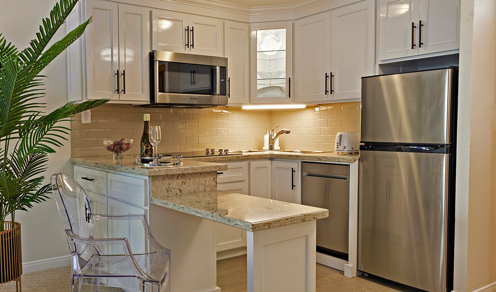 Modern kitchen with white cabinets, stainless steel appliances, beige granite countertops, a small peninsula with two glasses and a wine bottle, a clear chair, and a potted plant in the corner. Warm under-cabinet lighting.