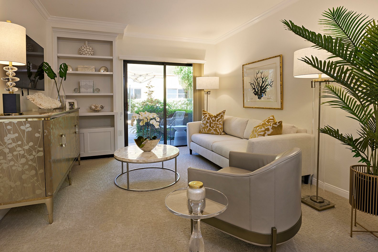 A modern living room with a white sofa, patterned pillows, a marble coffee table, armchair, floor lamp, potted plant, and glass doors opening to a patio. Neutral tones and gold accents create an elegant atmosphere.