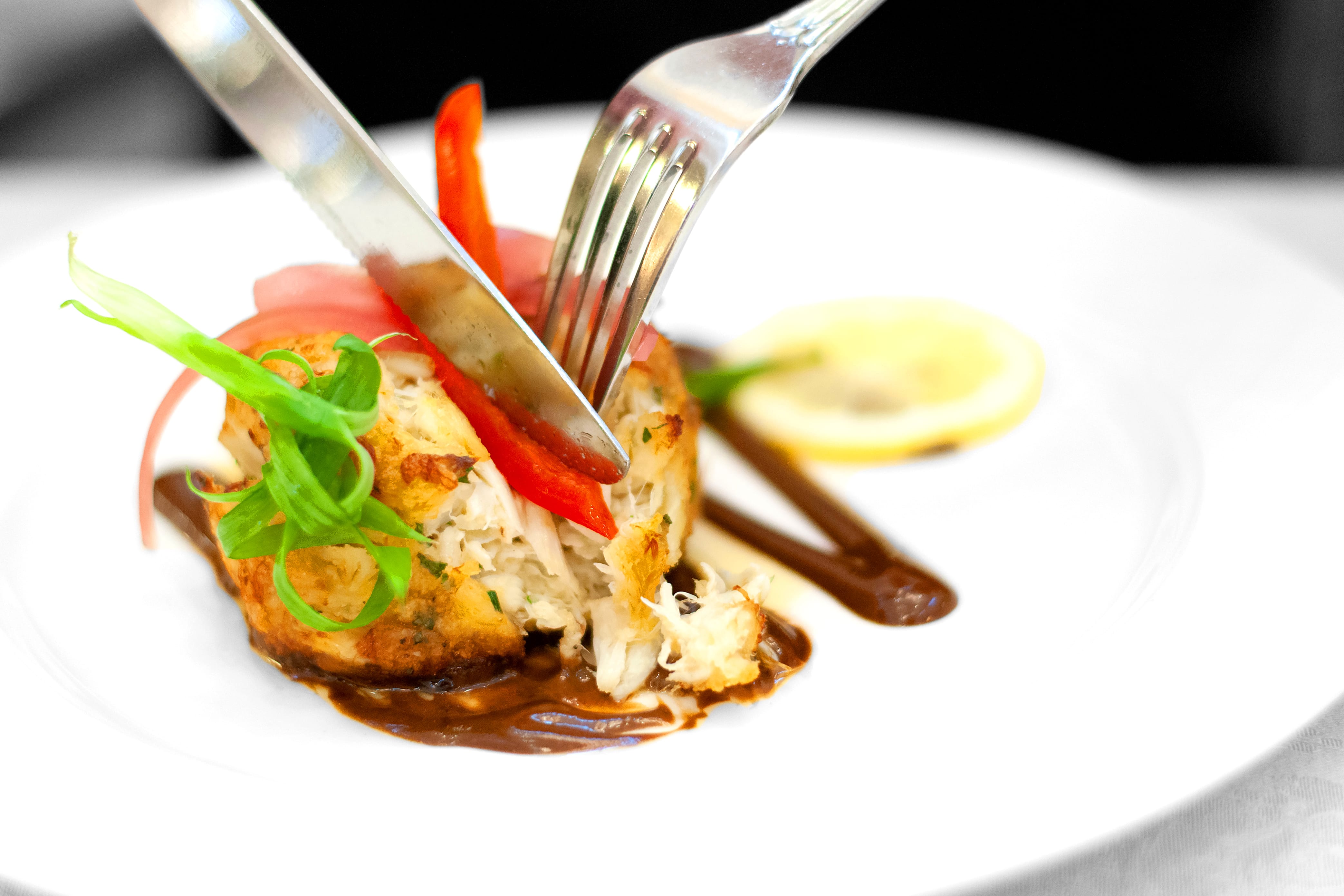 A fork and knife cut into a crab cake topped with red peppers, green onion, and pink pickled onions, served with brown sauce and a lemon slice on a white plate.