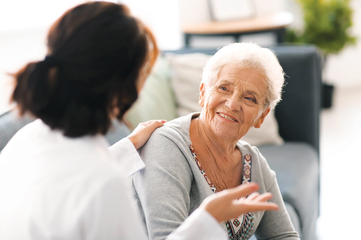 A smiling elderly woman sits on a couch, looking at and talking with a caregiver or healthcare professional, who rests a reassuring hand on her shoulder. The setting appears bright and homey.